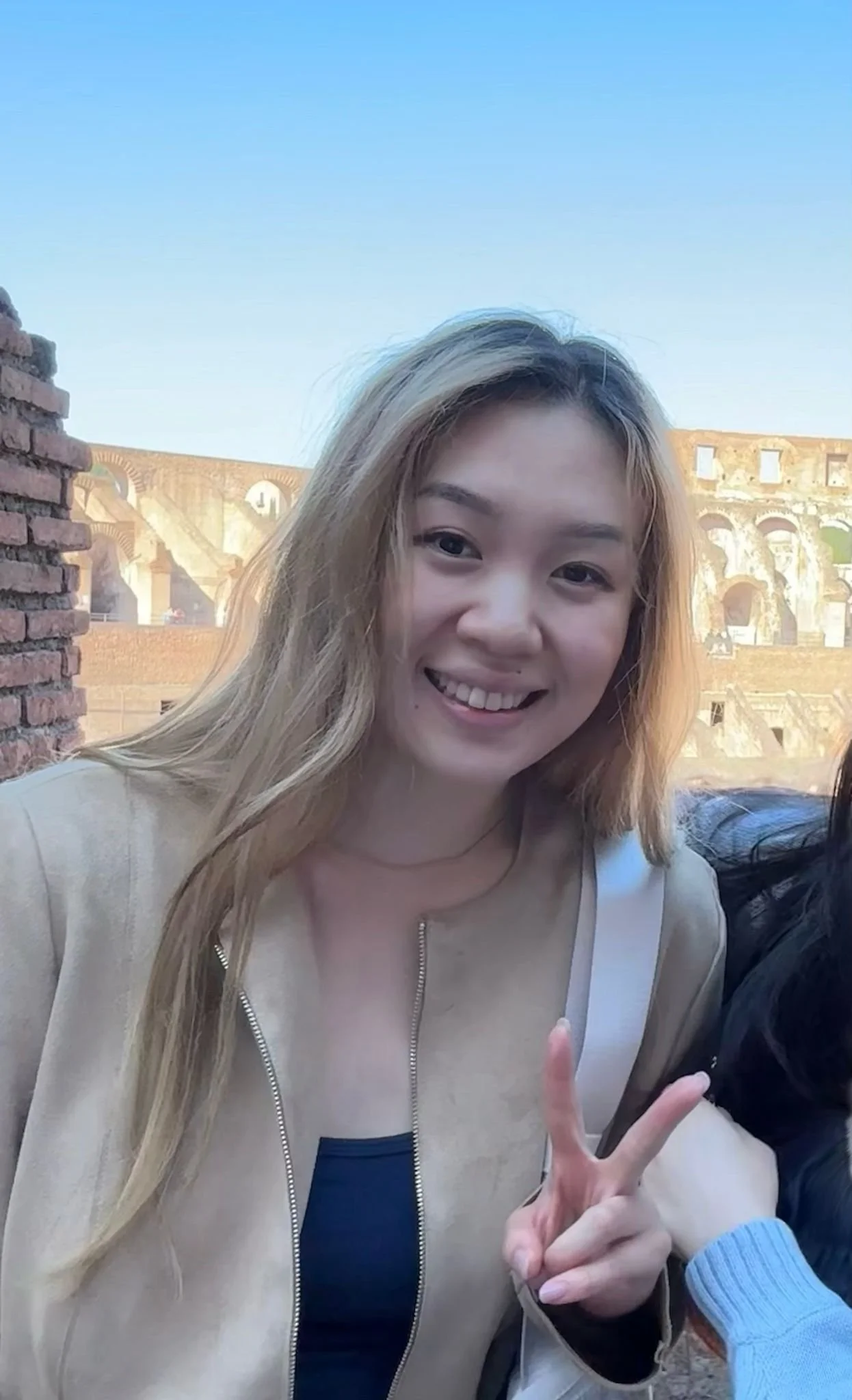 A smiling woman with blonde hair making a peace sign with her hand, in front of the ancient Roman Colosseum.