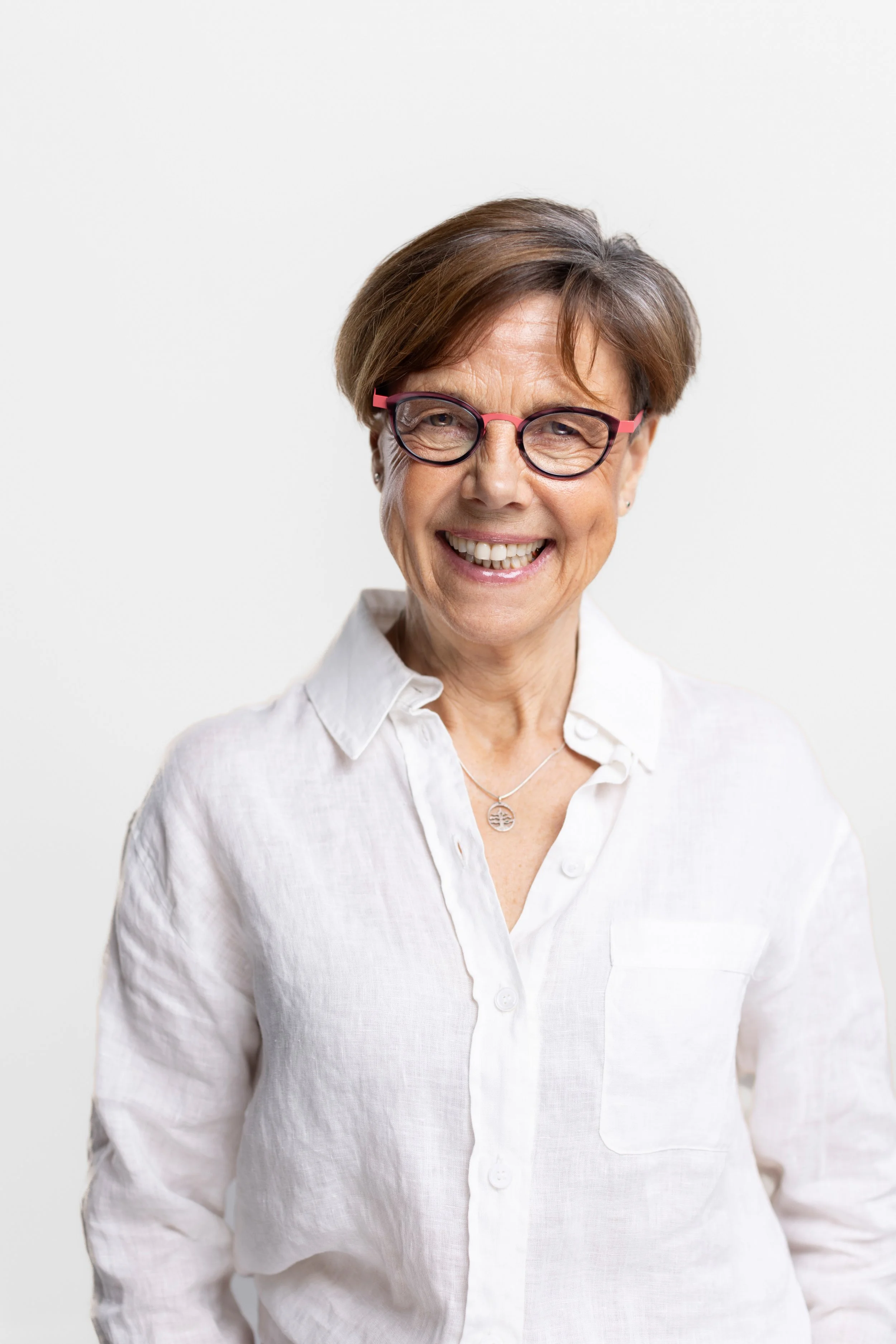 Smiling middle-aged woman with short brown hair, wearing glasses and a white button-up shirt, against a plain white background.