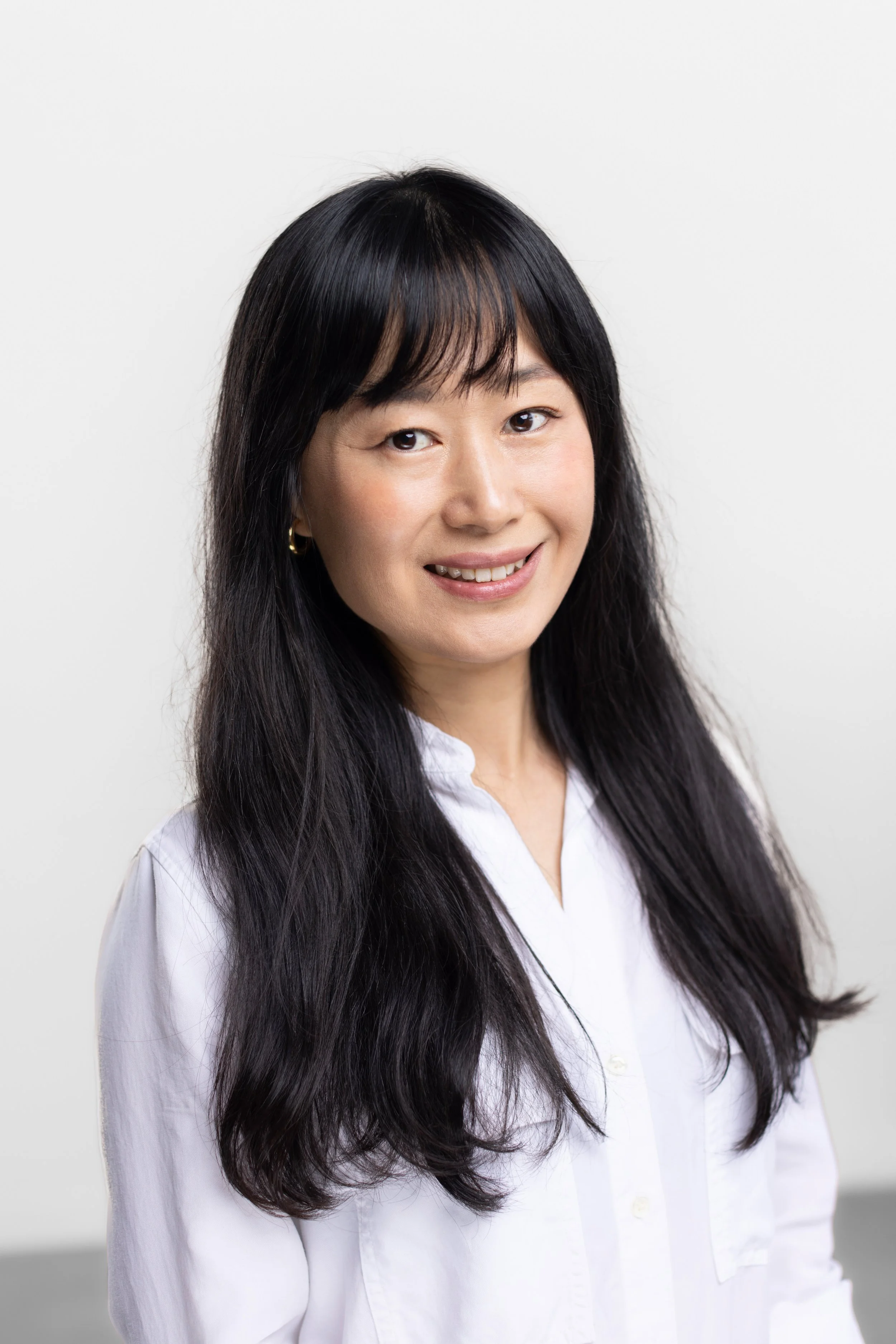 A woman with long black hair wearing a white blouse, smiling against a plain white background.