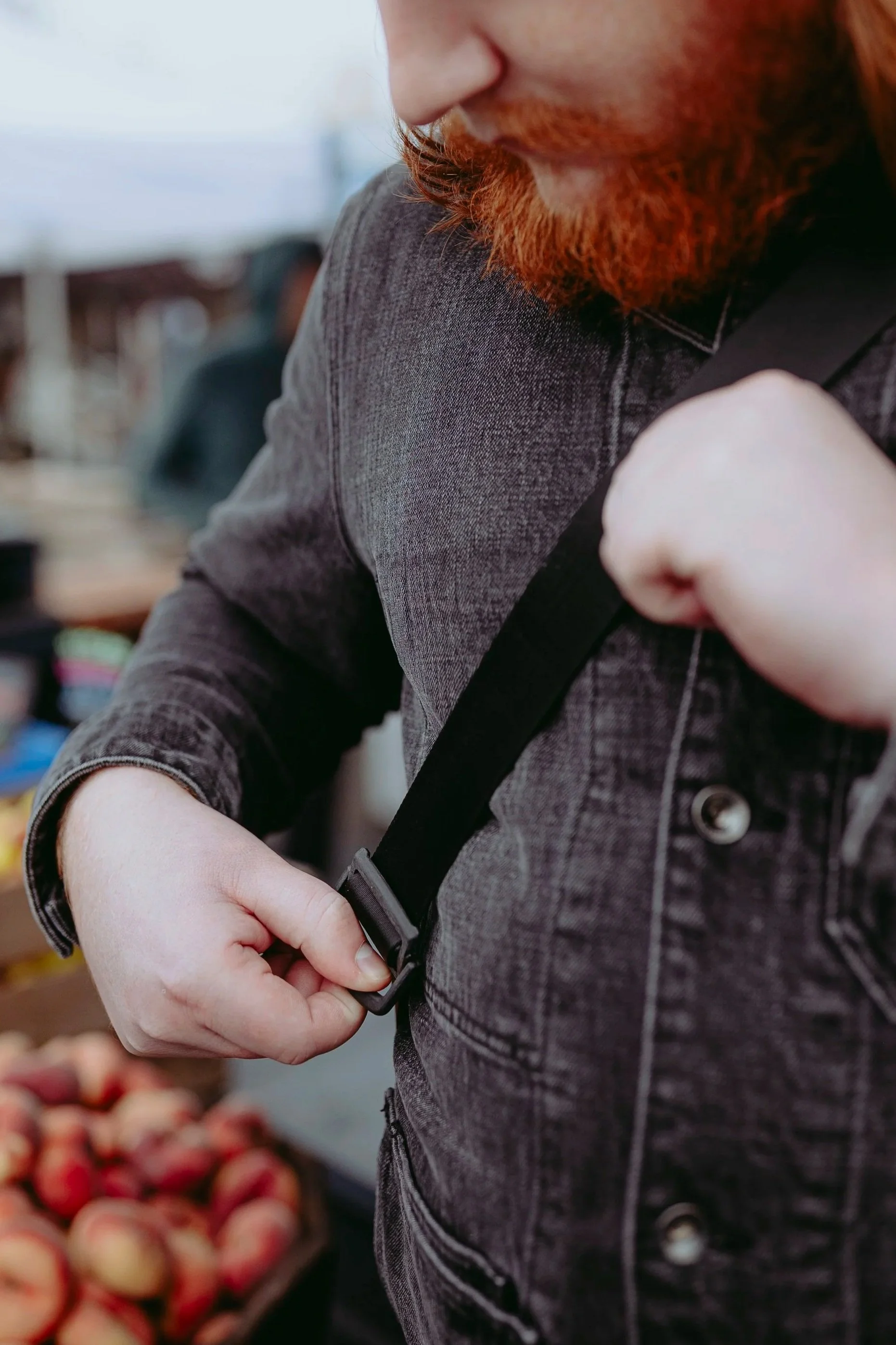 A person adjusting a black strap on their shoulder, wearing a dark gray denim jacket, at an outdoor market with peaches in the background.