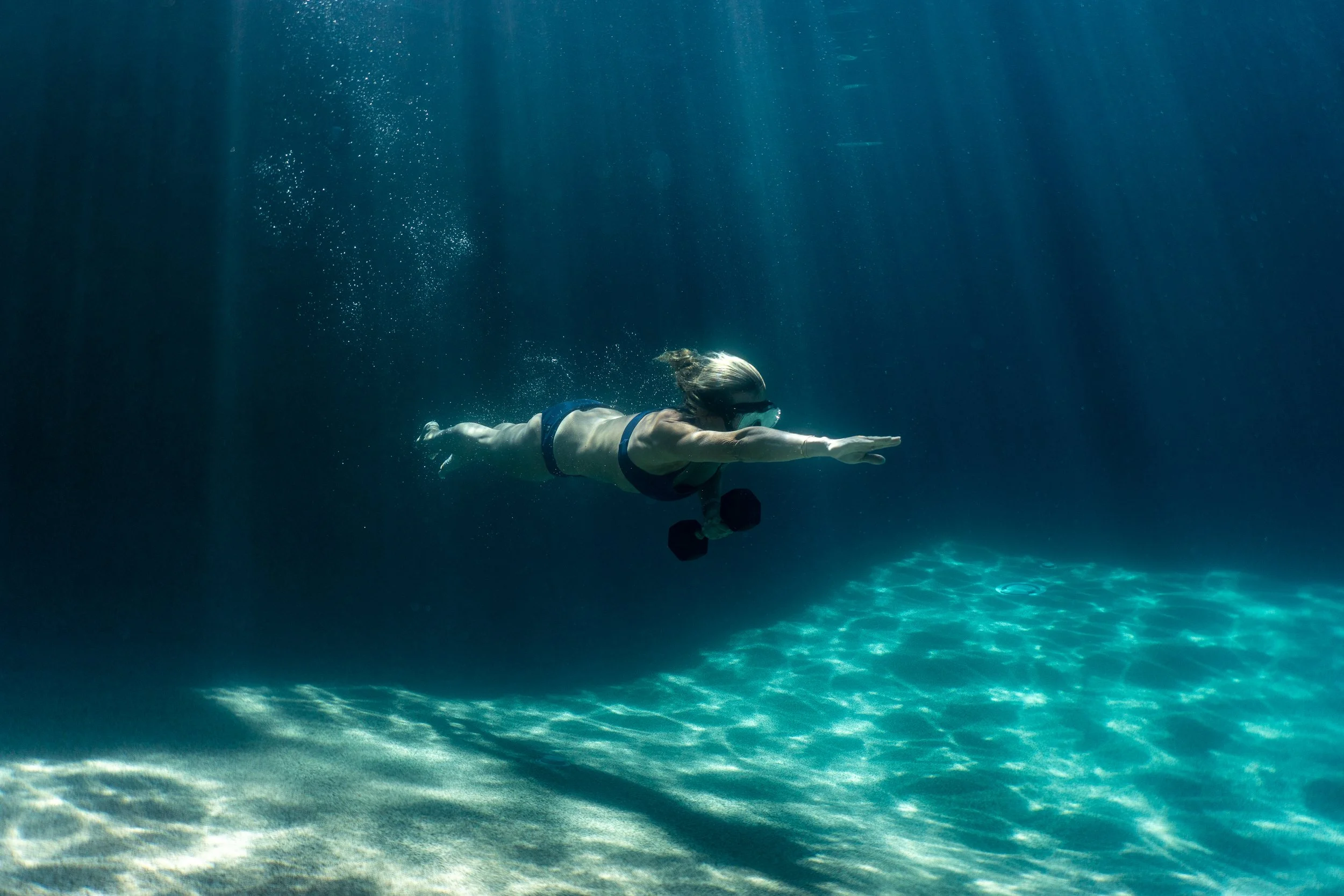 A woman swimming underwater wearing a black bikini, goggles, and holding dumbbells.