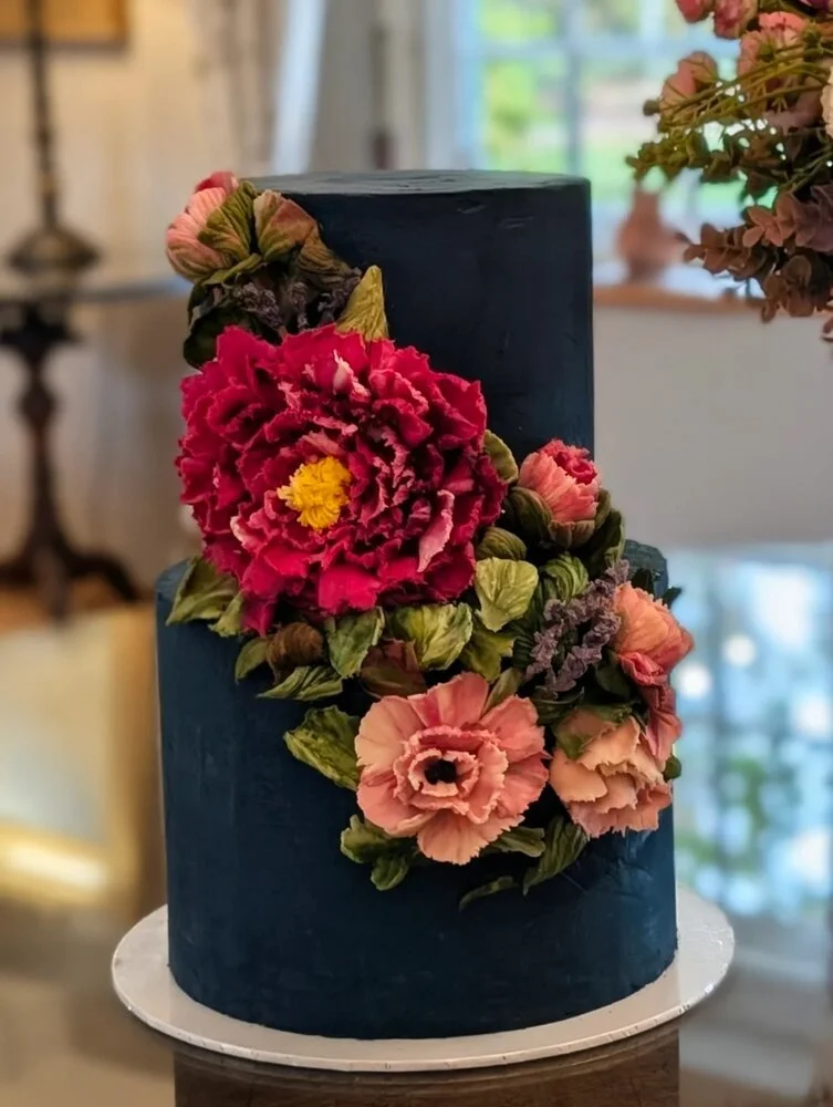 A two-tiered dark blue wedding cake, decorated with bold pink and red peonies hand-sculpted in Belgian white chocolate.  