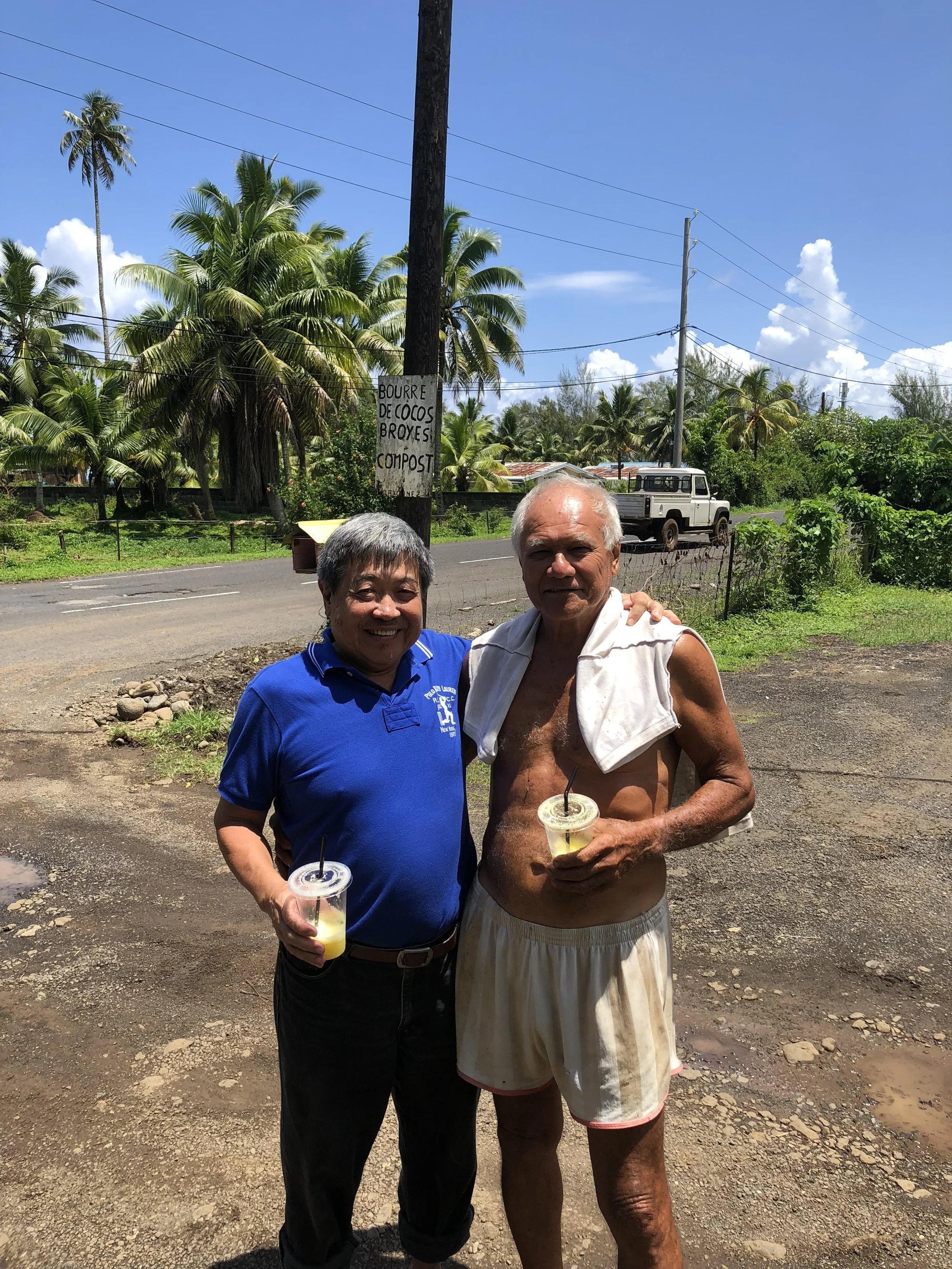 Two men standing outdoors smiling, holding drinks, with tropical palm trees and a sign on a pole behind them. One man is wearing a blue polo shirt, and the other is shirtless with a towel over his shoulders.