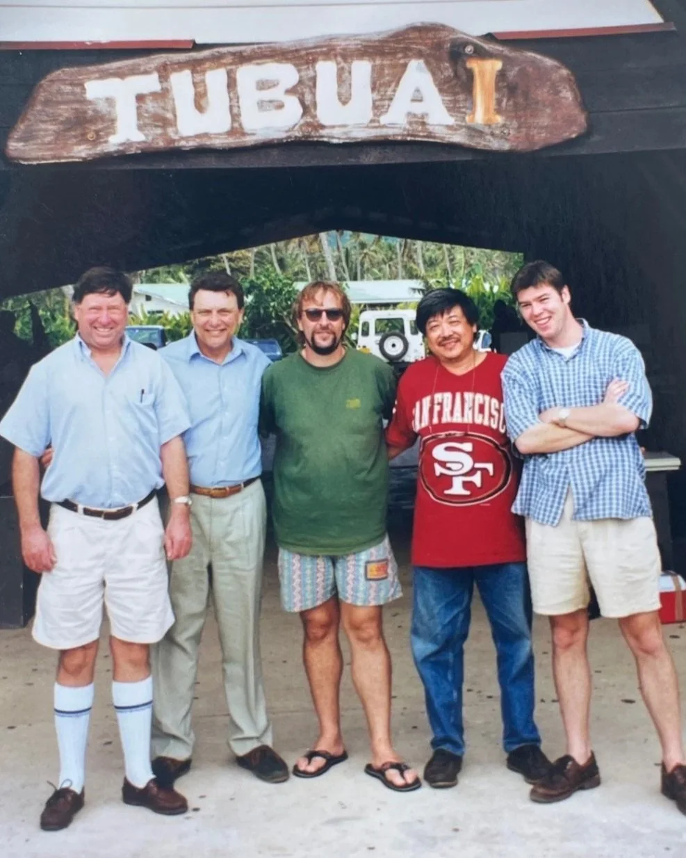 Group of five men standing outdoors in front of a large wooden sign that reads "TUBUA" with a small "1" on it. The men are smiling and dressed casually, with some in shorts and others in shirts and pants. There are trees and a vehicle in the background.