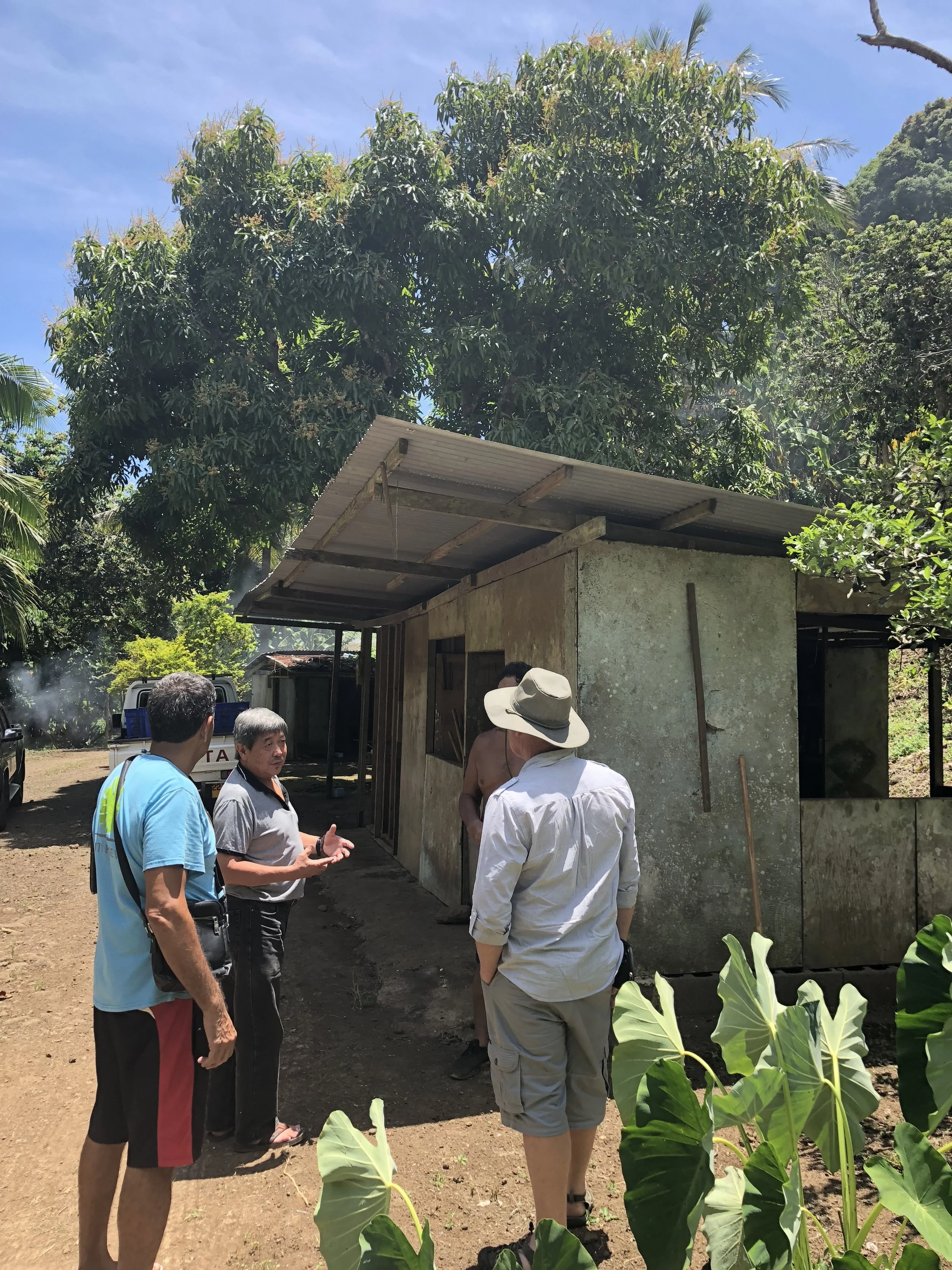 A group of four men talking outside a small building in a lush, tropical environment under a partly cloudy sky.