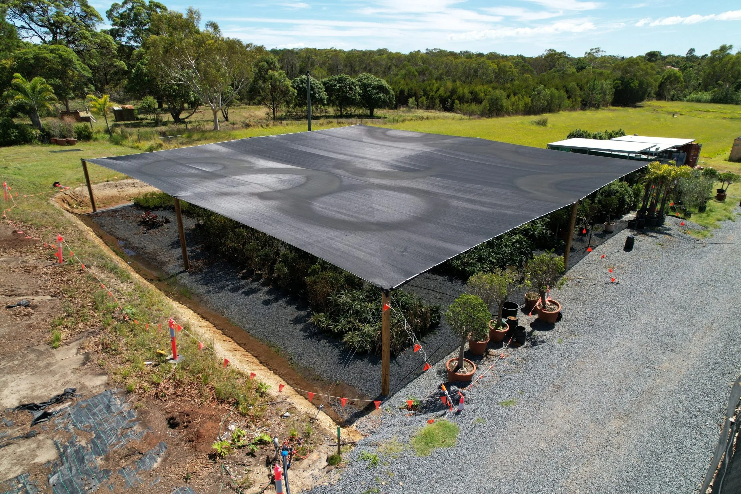 A large outdoor shade cloth supported by wooden posts covers a garden and gravel pathway area in a rural setting, with trees and open fields in the background.