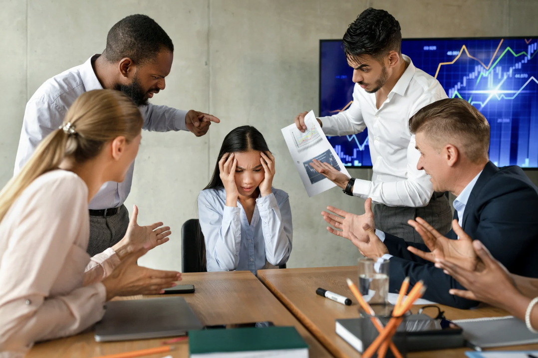 A woman at a business meeting looks stressed and overwhelmed as colleagues argue around her, with a large screen displaying stock market graphs in the background.