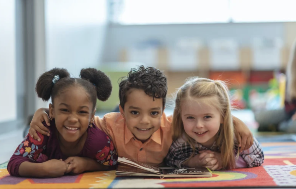 Three young children lying on a colorful classroom carpet, smiling, with an open book in front of them.