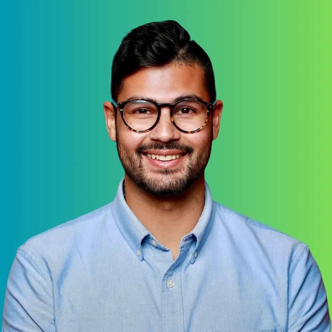 A young man with dark hair, glasses, and a beard, smiling in front of a green and blue gradient background, wearing a light blue collared shirt.