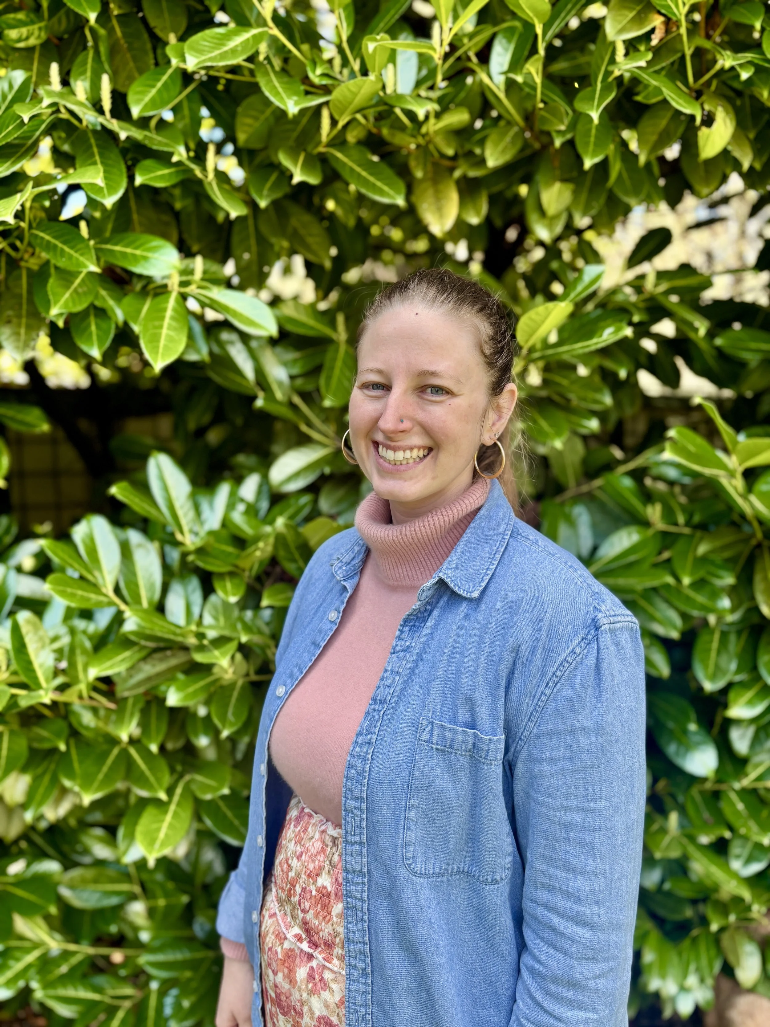 A woman with blonde hair tied back, wearing a pink turtleneck, a floral dress, and a blue denim shirt, standing outdoors in front of green leafy bushes, smiling at the camera.