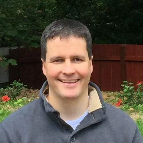 A man with short dark hair smiling, outdoors with trees, flowers, and a red wooden fence in the background.