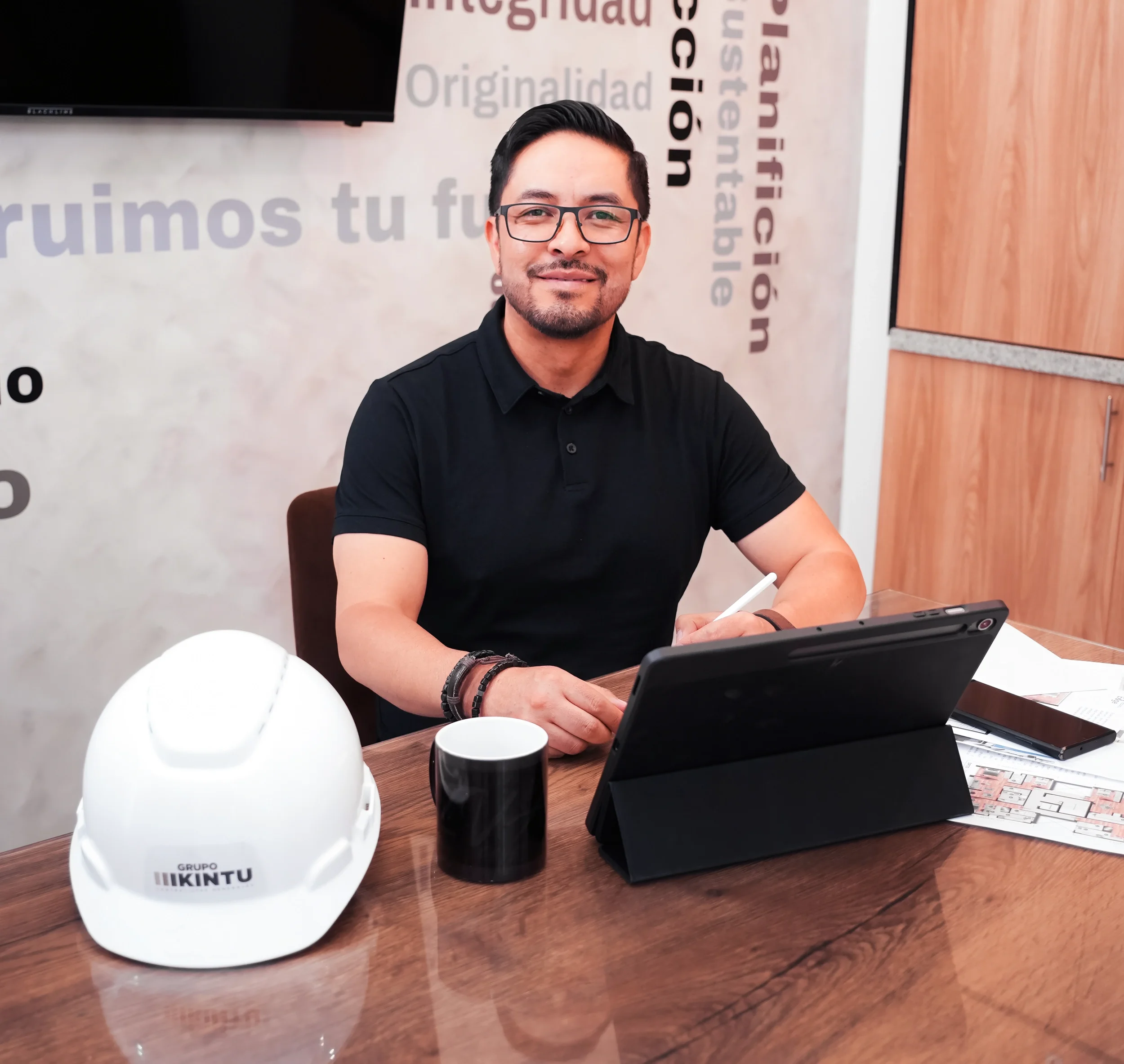 Hombre sentado en una mesa de oficina, sonriendo, con escritorio que tiene cascos de seguridad, una taza, un tablet y documentos, fondo con palabras relacionadas con innovación y originalidad.