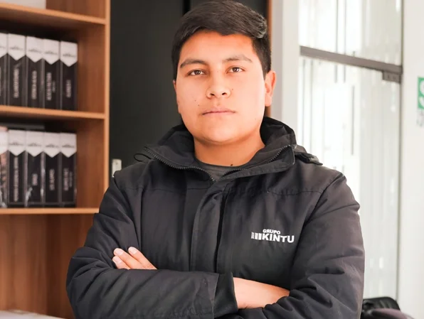 Joven con chaqueta negra con el logotipo 'Grupo KINTU' de pie con los brazos cruzados, en un entorno de oficina o espacio de trabajo.