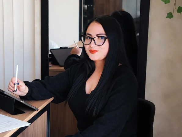 Mujer con gafas negras y cabello largo sentado en un escritorio, sosteniendo un bolígrafo y con una tableta frente a ella, en un ambiente de oficina.