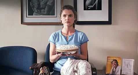 A scene from the movie The Waitress. A woman sitting on a blue chair holding a cake with white frosting and decorations, in a room with framed photos on the wall.