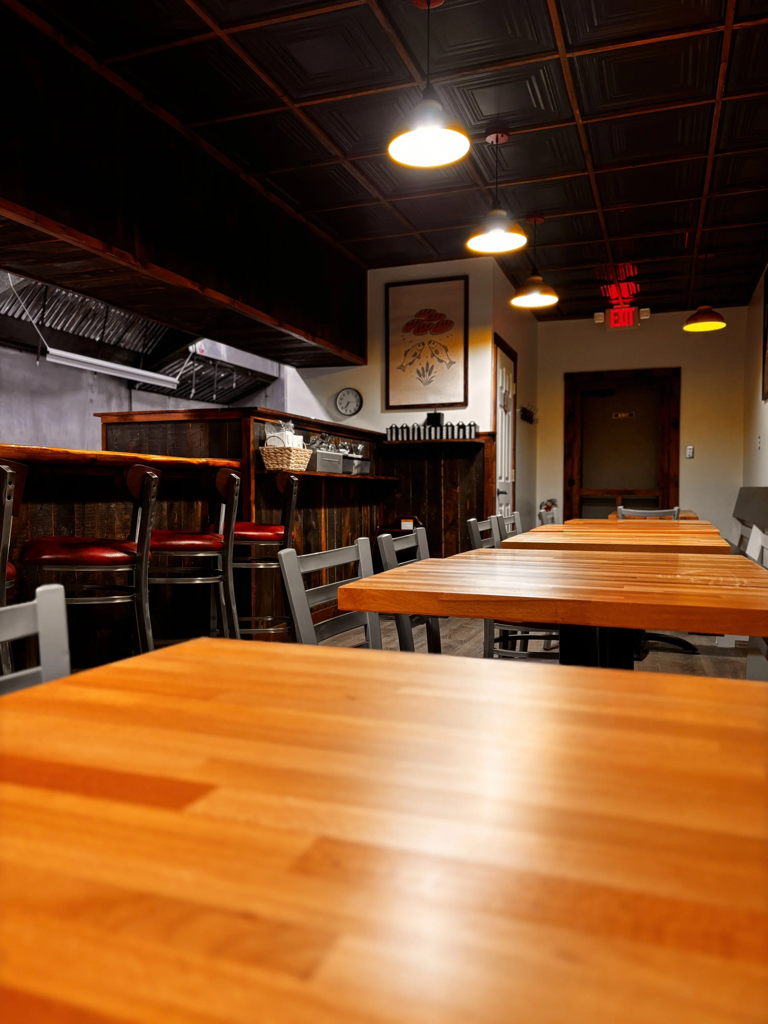 Empty restaurant dining area with wooden tables and chairs, warm lighting, and a bar counter in the background.