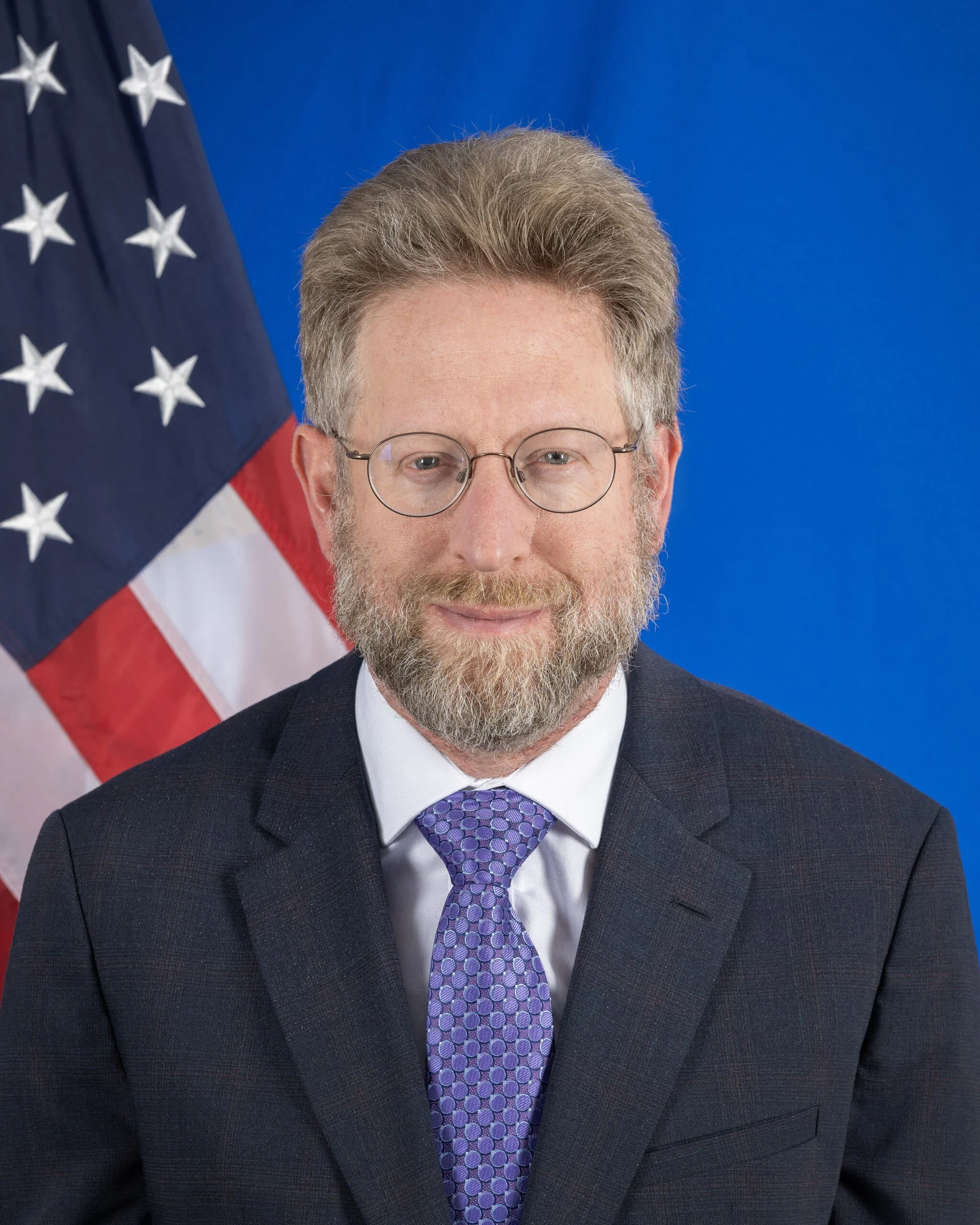 A man in a dark suit, white shirt, and purple tie, wearing glasses, standing in front of an American flag and a blue background.