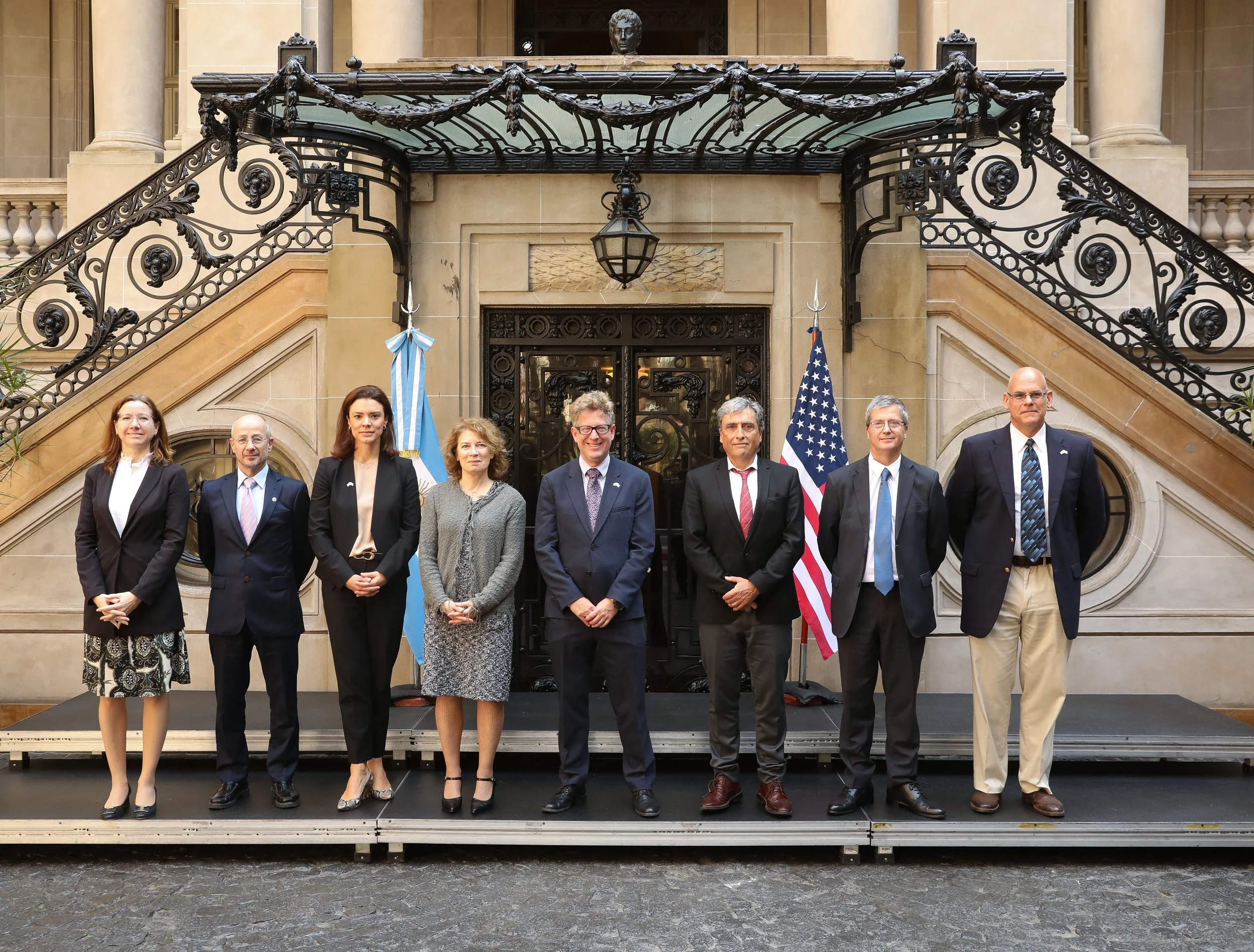 A group of nine officials standing on a platform in front of a building with ornate wrought-iron stairs and a bronze bust on top. Two flags are visible: the American flag and a blue and white striped flag.