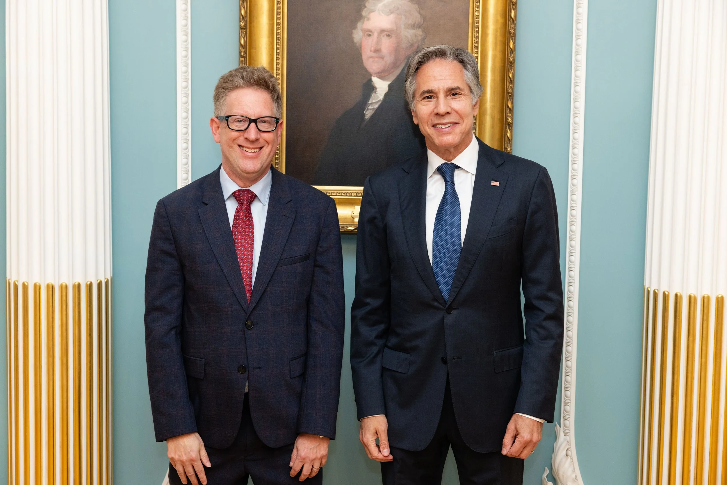 Two men in suits standing in front of a framed portrait of a historical figure, in a room with classical decor.