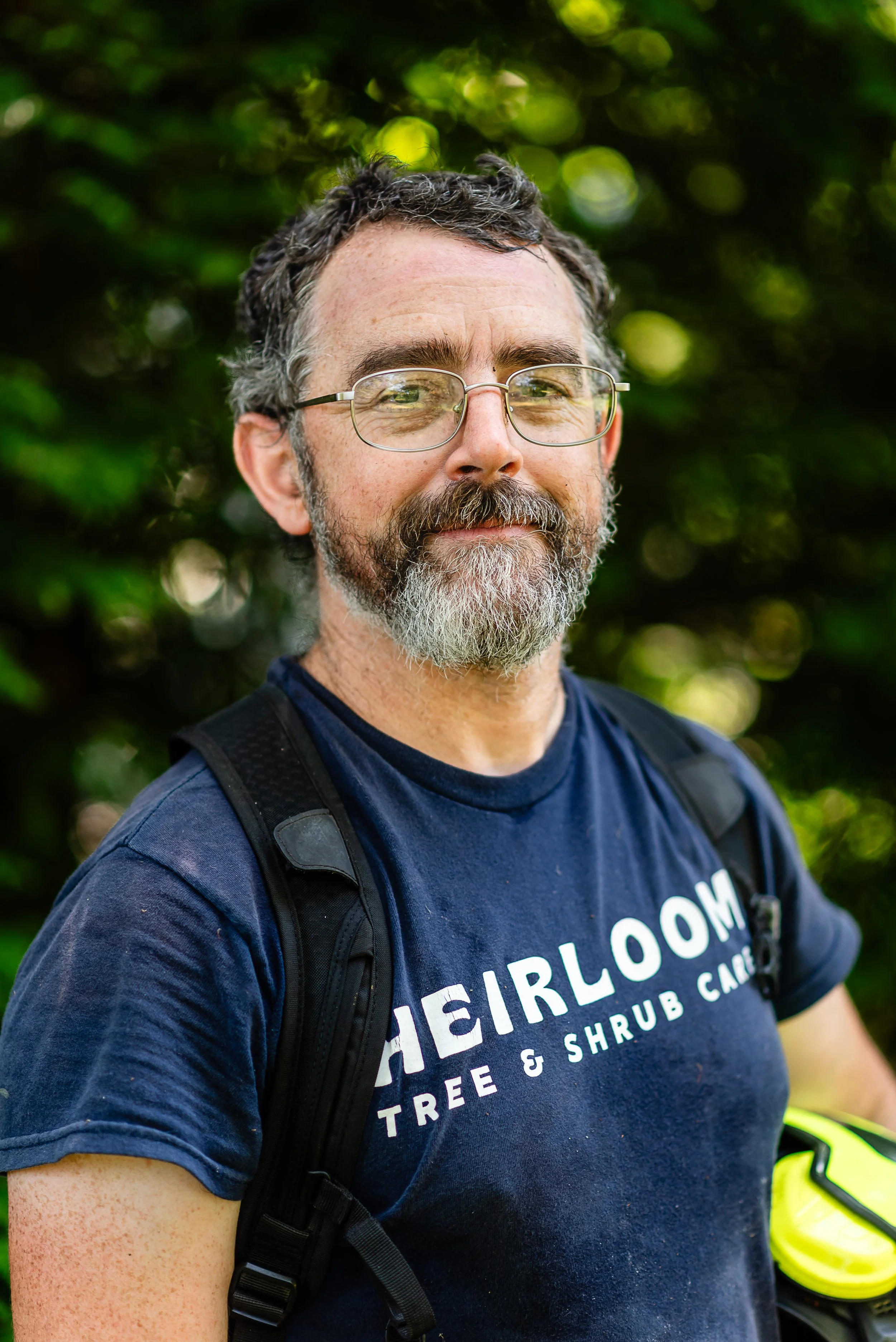 A middle-aged man with glasses and a beard, wearing a navy blue T-shirt and a backpack, standing outdoors with a background of blurred green foliage.