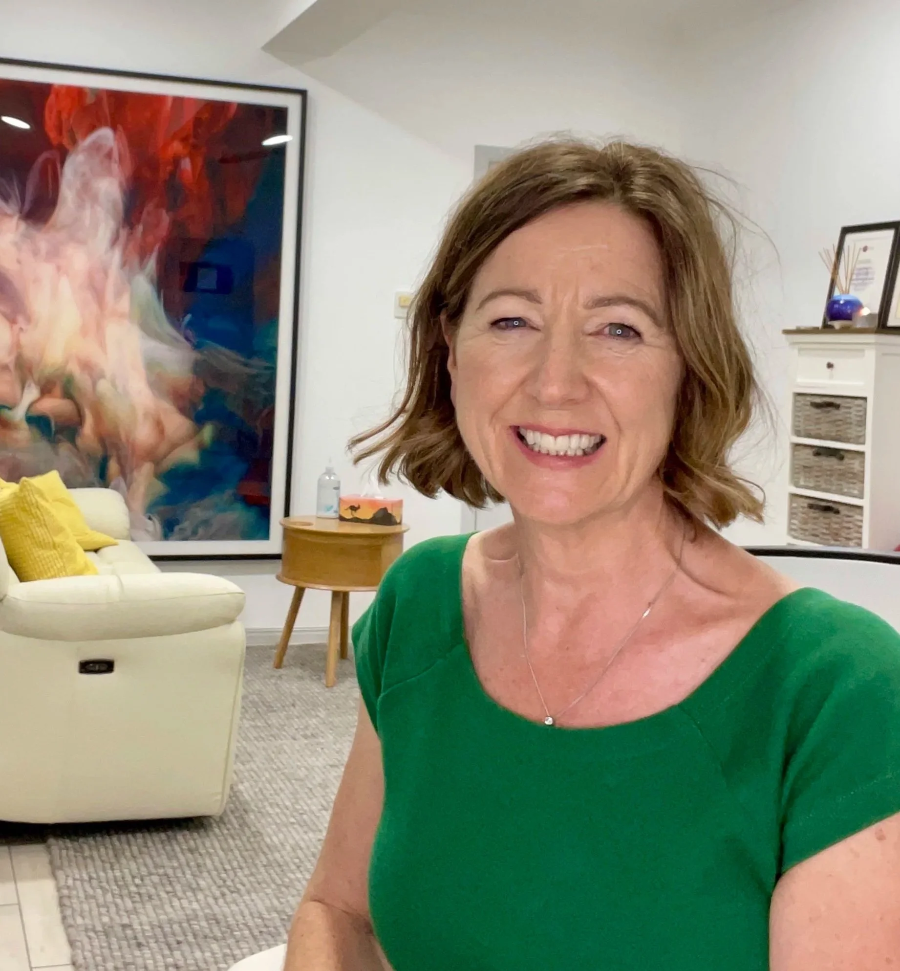 Close-up of a woman smiling, wearing a green top, in a modern living room with artwork and furniture.