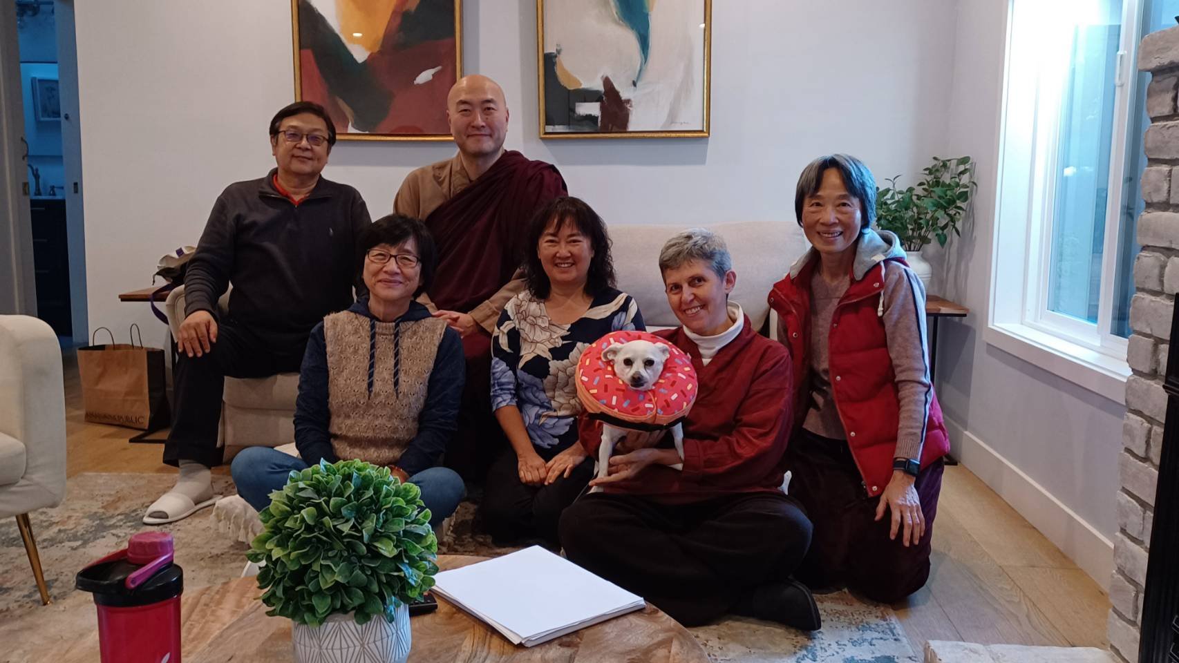 A group of seven people and a dog, sitting and standing in a cozy living room. The dog is wearing a donut-shaped collar. There are abstract paintings on the wall, a potted plant, and a window with natural light. The group is smiling, and there is a table with a plant, a notebook, and a pink water bottle in the foreground.