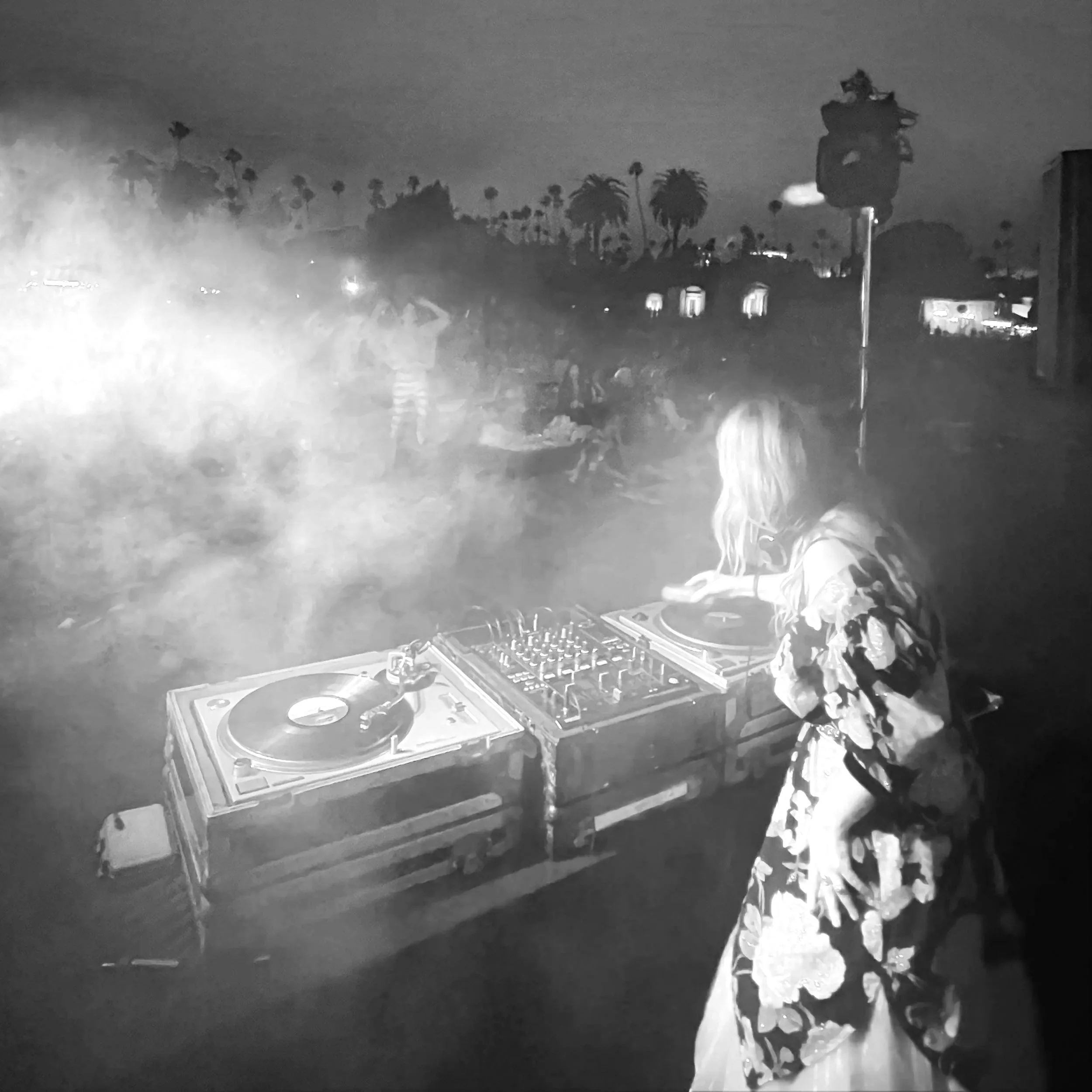 A female DJ with long hair in a floral dress performs at an outdoor concert at dusk Cinespia Los Angeles Pride Hollywood Forever Cemetary, with a crowd of people and palm trees in the background.