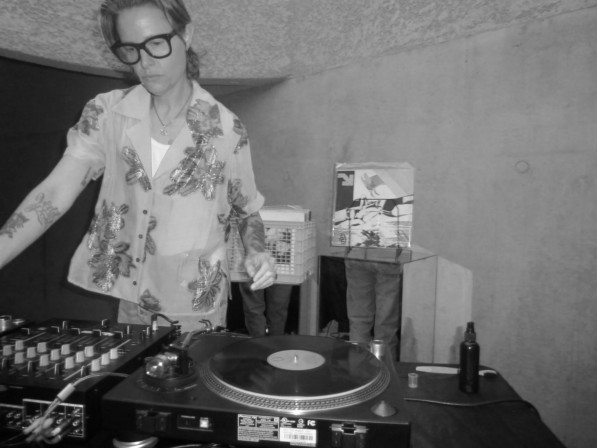 A woman DJ wearing glasses, a pink shirt with butterfly patterns, and tattoos, mixing music on DJ equipment in a dimly lit room.