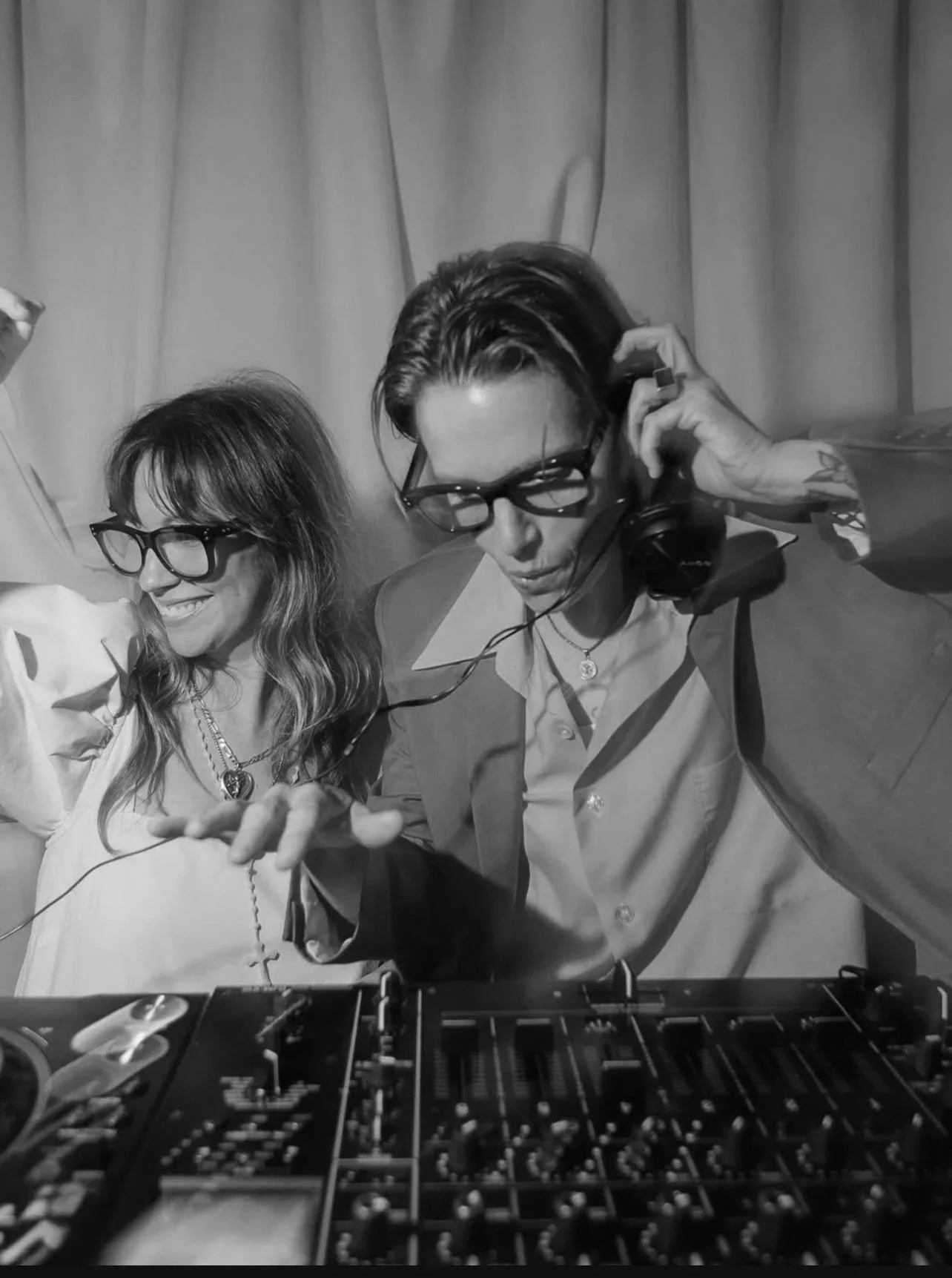 DJs Dash and sera, one with long wavy hair and the other with short hair, are working as DJs in a room with pink curtains. They are using DJ equipment, and one woman is listening to music with headphones while the other is smiling and gesturing.