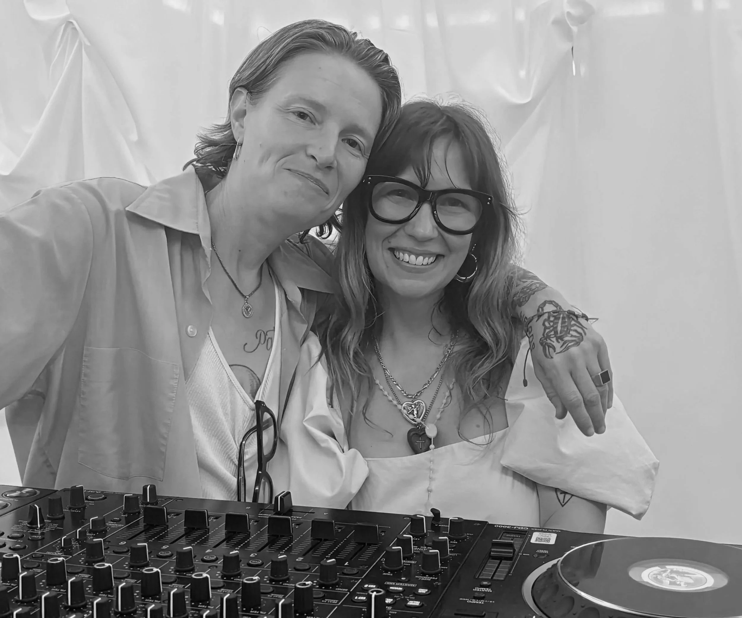 Two women posing together next to DJ equipment, one with short hair and the other with long, wavy hair and glasses, both smiling and close to each other