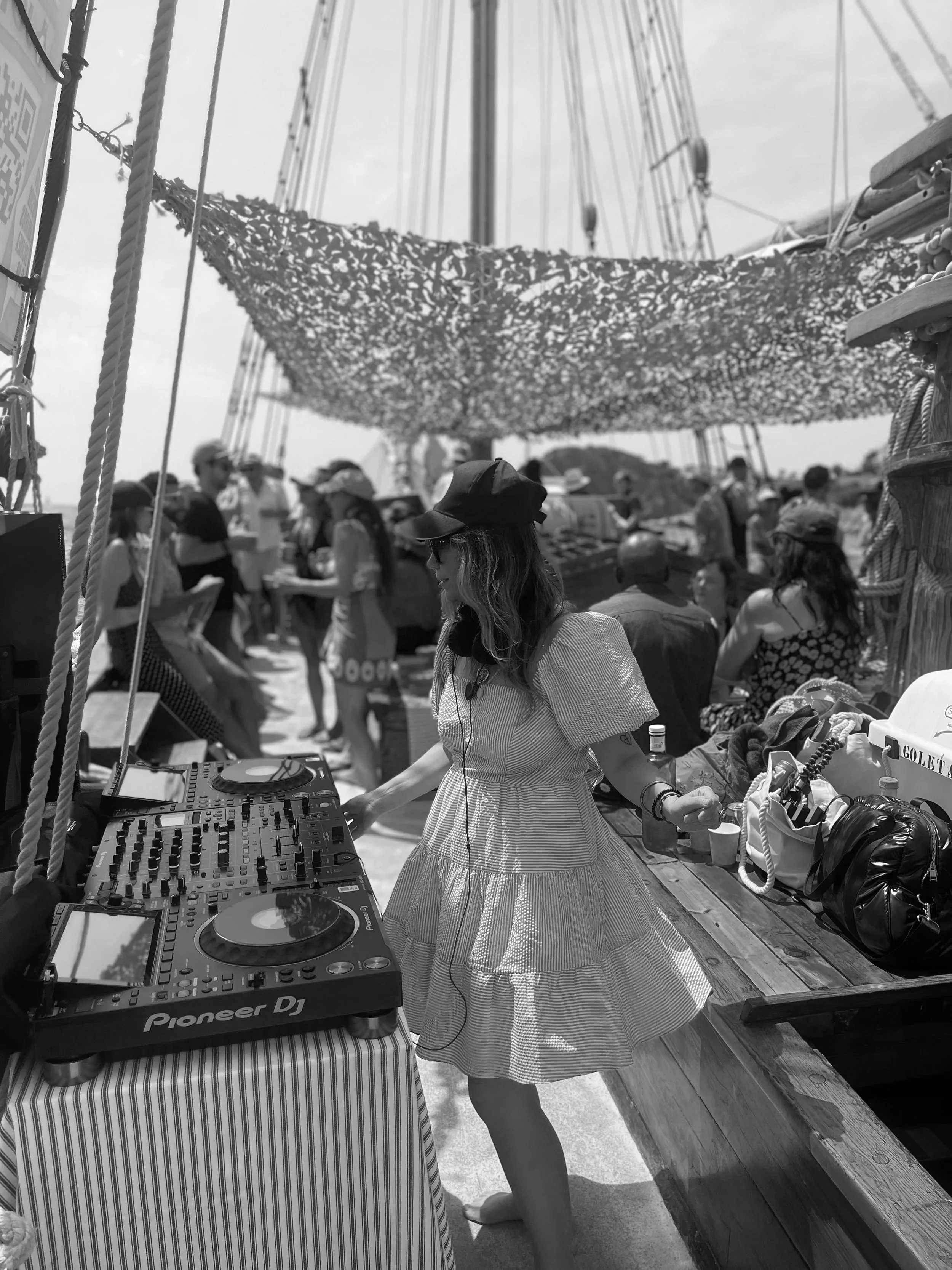 A woman in a blue and white striped dress and black hat DJing at a beach party on a boat, with a crowd of people dancing and socializing in the background.