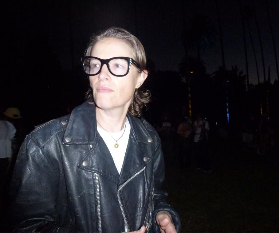 dash lippman (they) with short brown hair and glasses wearing a black leather jacket, standing outdoors at night with palm trees and people in the background.