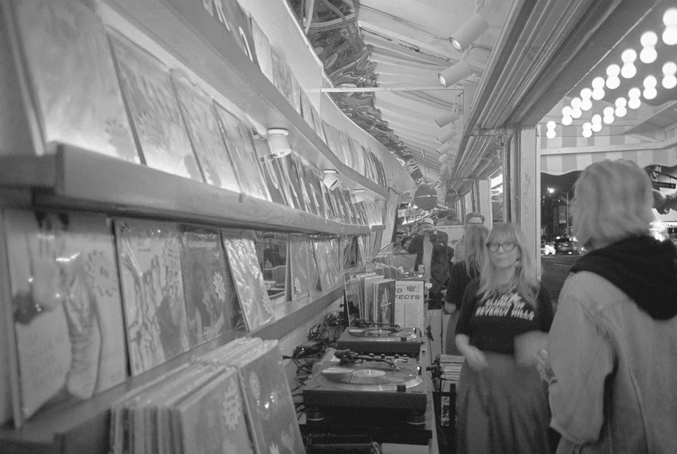 People browsing and shopping for records at an outdoor vinyl record store at night, illuminated by warm lighting and string lights. Sunset blvd brand party collab with searchlight across street from chateau marmont