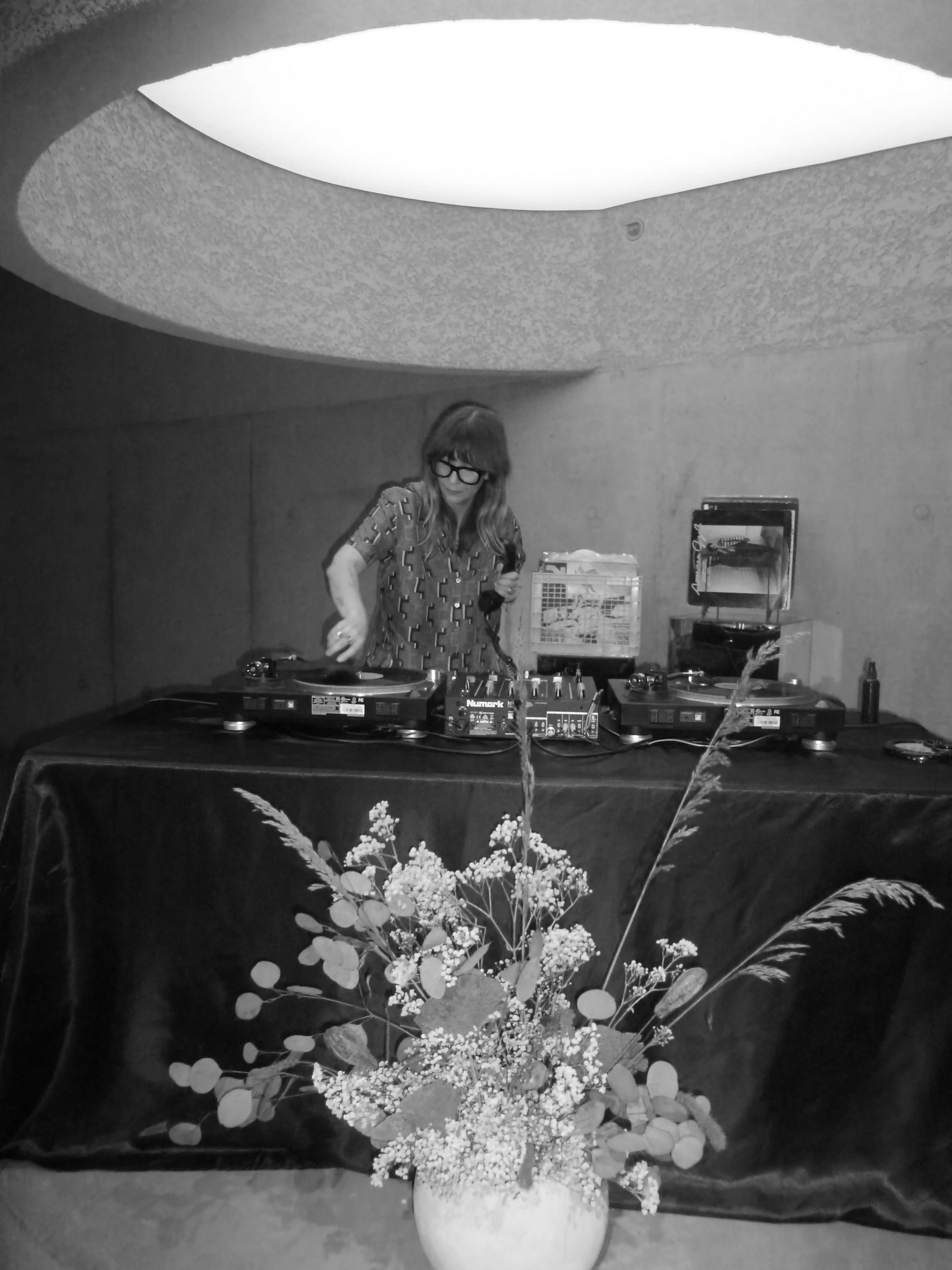 A woman DJ wearing a red patterned shirt and glasses is performing with turntables at an indoor event, decorated with a large pink floral arrangement in the foreground.
