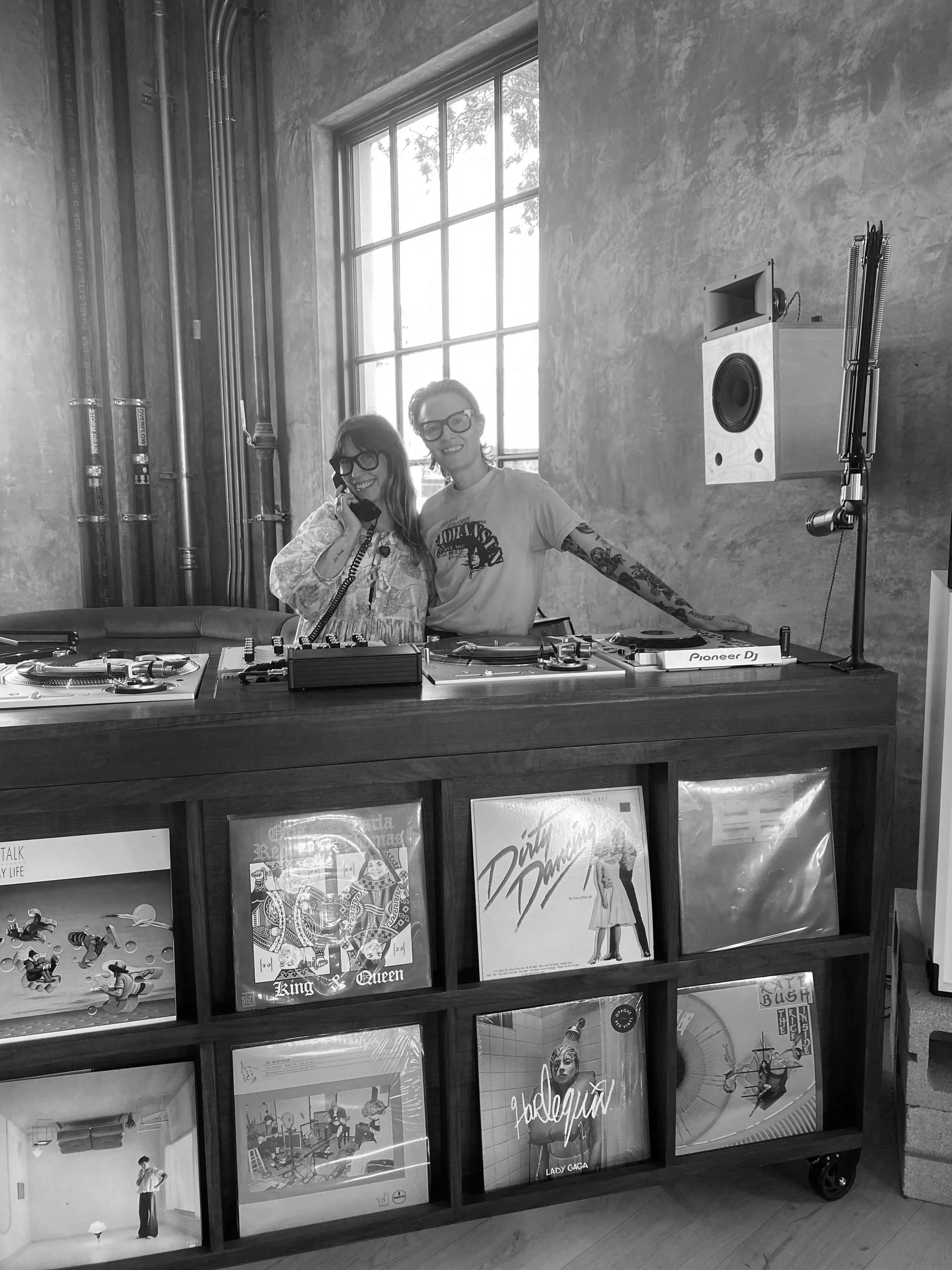 Two women wearing glasses smiling behind a DJ booth with vinyl records displayed in a wooden shelf in front. One woman is talking on a phone, and there is a large window behind them with some greenery visible outside.