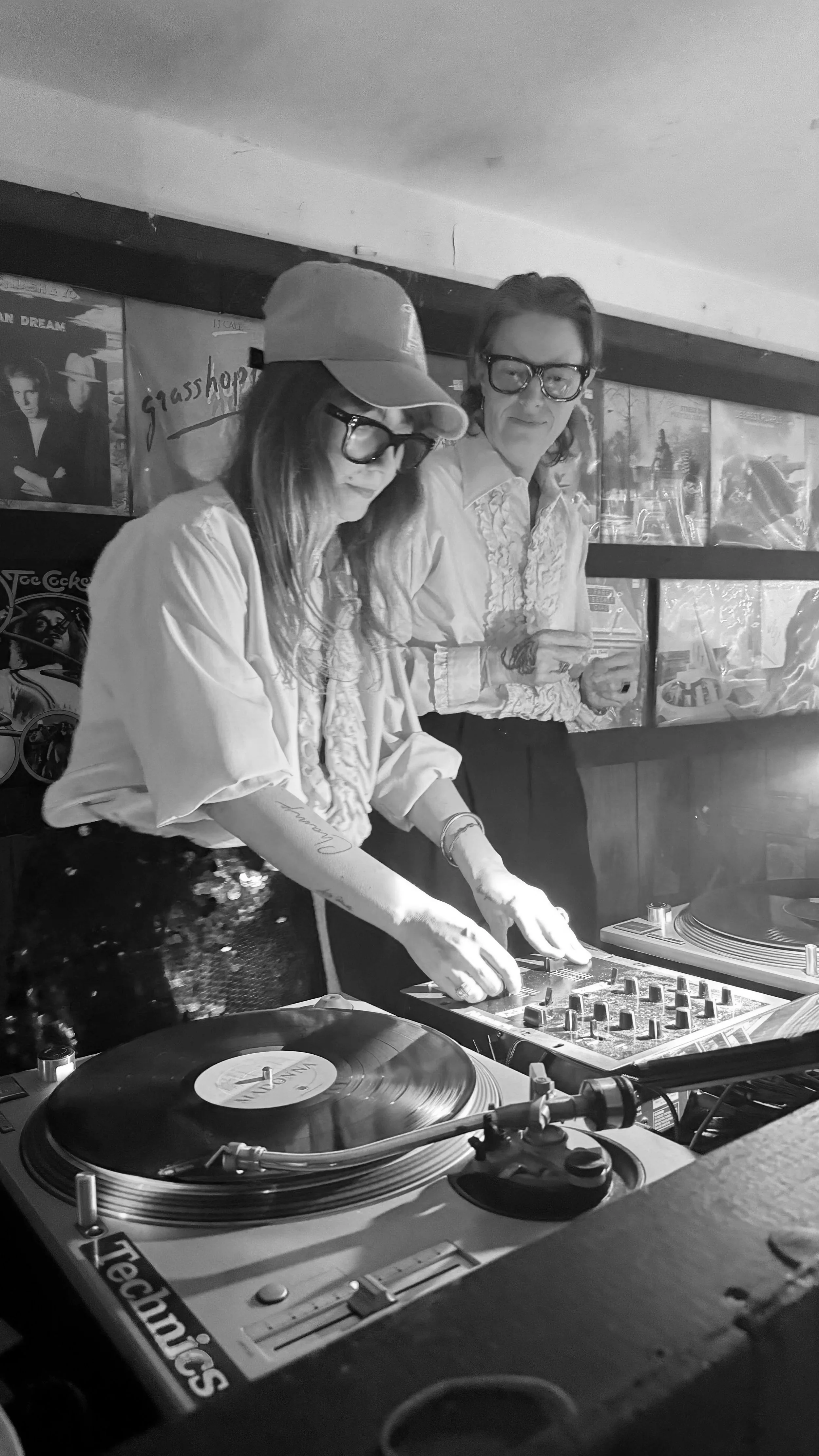 Two women DJing at a club, one wearing a pink blouse, glasses, and a cap, the other wearing a white blouse and glasses, with posters on the wall behind them.