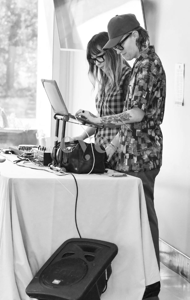 Two young people with glasses and tattoos using a laptop at a DJ setup with mixer and speaker in a well-lit room.