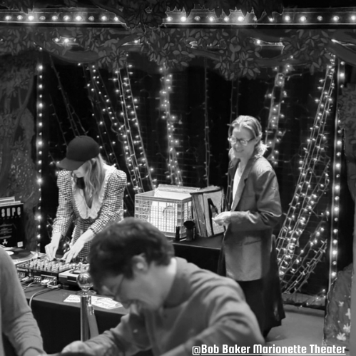 Two women are at a DJ booth at Bob Baker Marionette Theater playing music for an all ages dance party including puppets, with colorful string lights and decorations in the background, with a man in the foreground looking down.