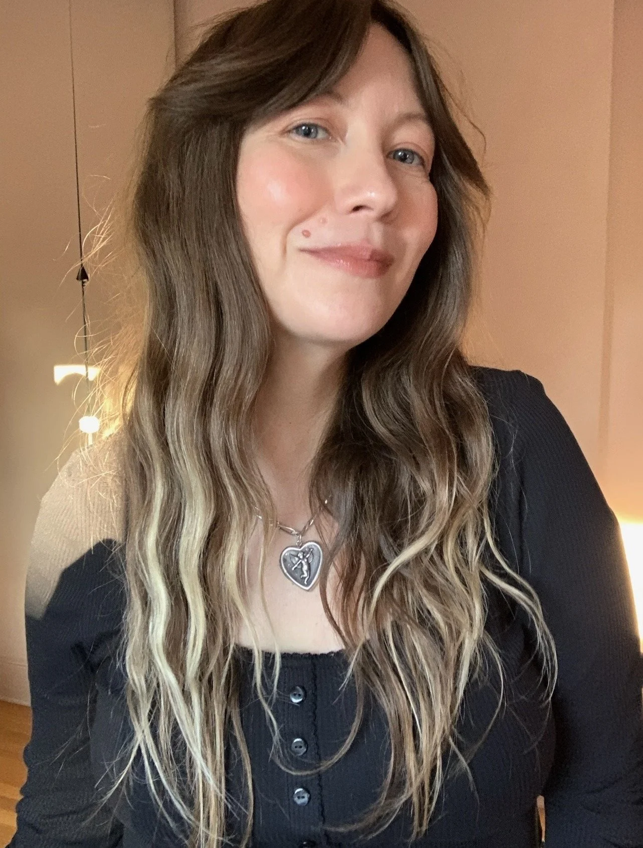 sera sloane with long, wavy light brown hair with blonde highlights, wearing a black top, a necklace with a pendant, and smiling slightly. The background includes beige and warm lighting.