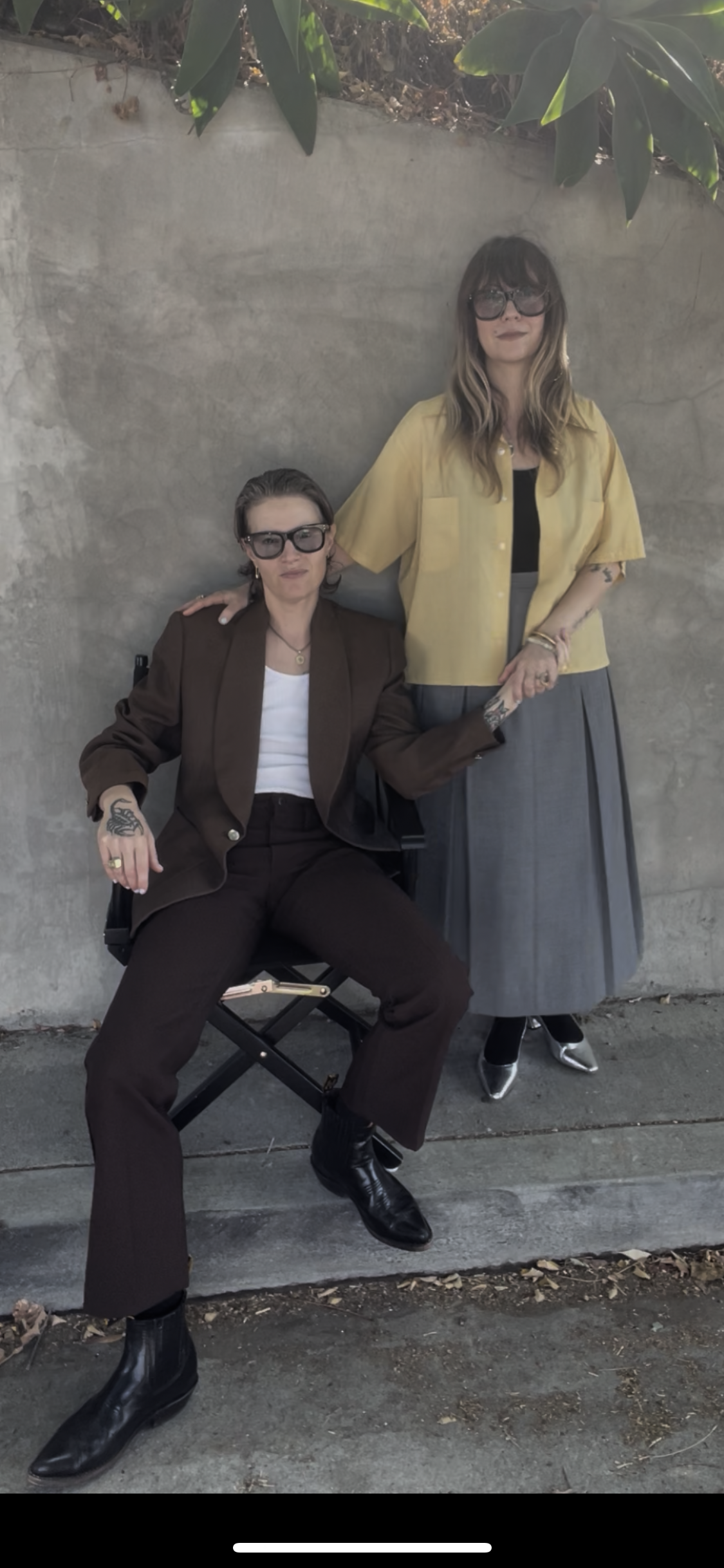 Two women with tattoos and glasses pose in front of a concrete wall, one sitting on a director's chair with a brown suit and the other standing with a yellow shirt and a gray skirt.