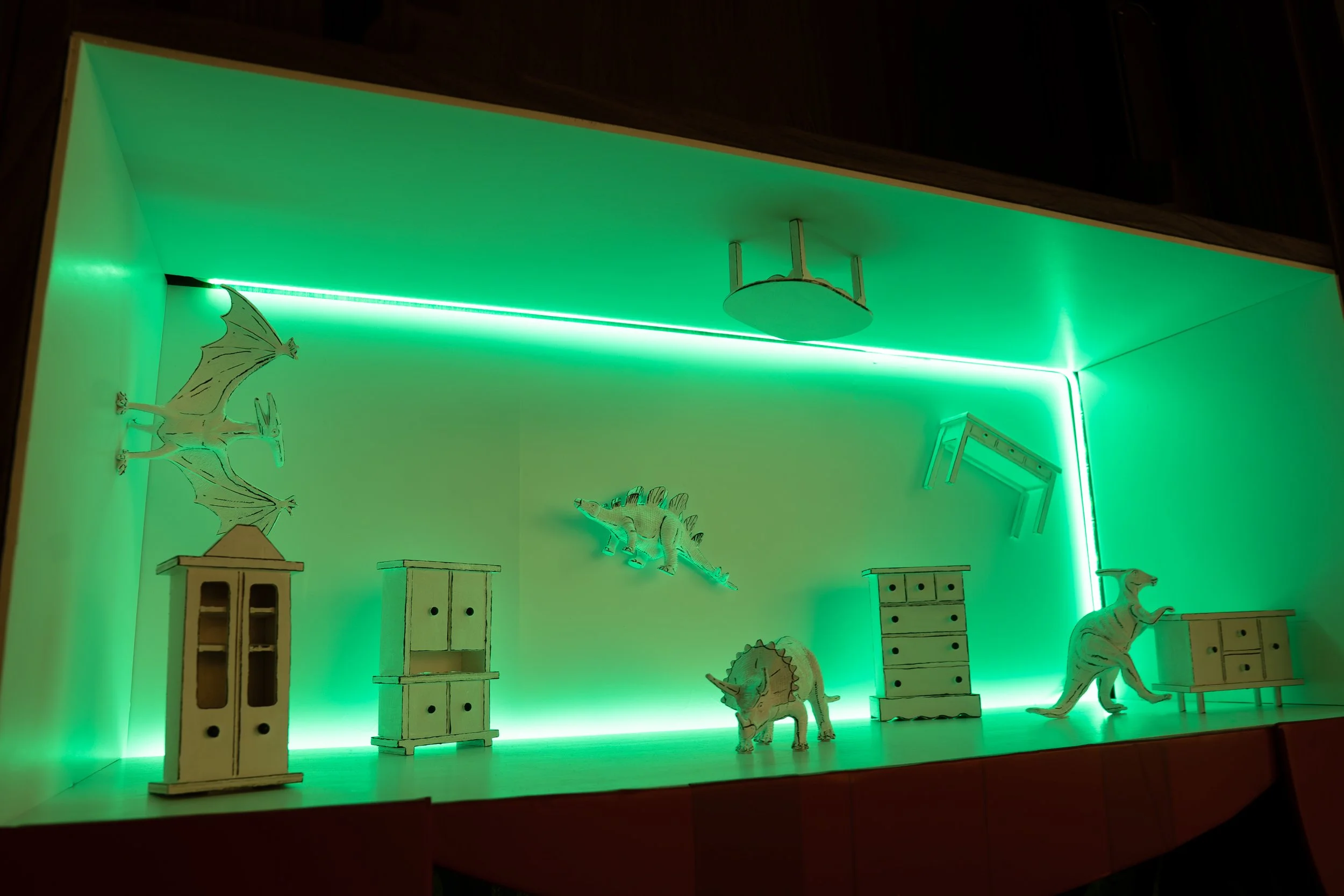 A green room with tiny surreal furniture and and dinosaurs attached to the walls and ceiling.