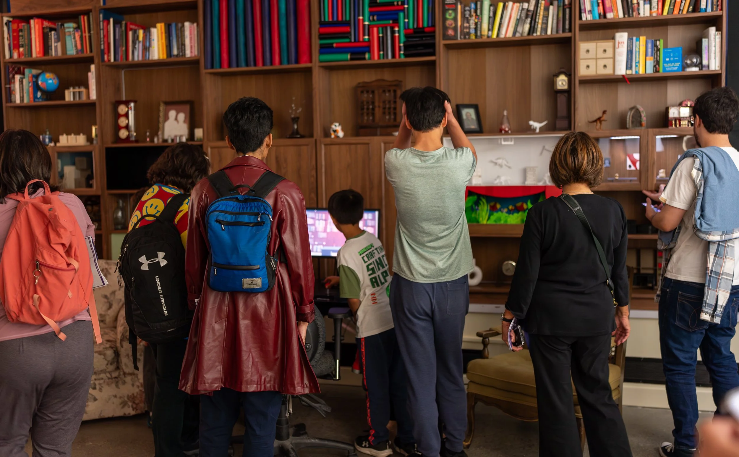 A crowd of people observing a video game and very full bookshelf
