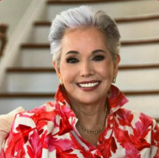 A woman with short, gray hair smiling, wearing a red floral blouse and necklace, standing in front of wooden stairs.
