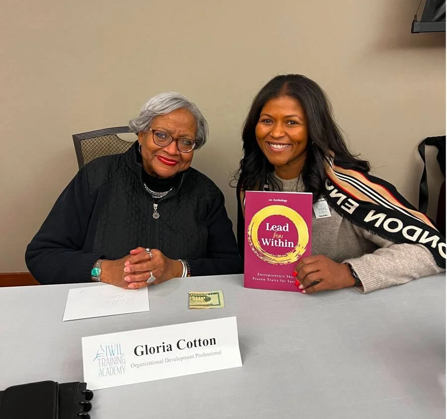 Two women sit at a table; one is elderly with gray hair and glasses, and the other is younger with black hair. The younger woman holds a pink book titled 'Lead from Within,' and both are smiling. There is a name card on the table with the name 'Gloria Cotton' and the title 'Organizational Development Professional,' along with a dollar bill and some papers.