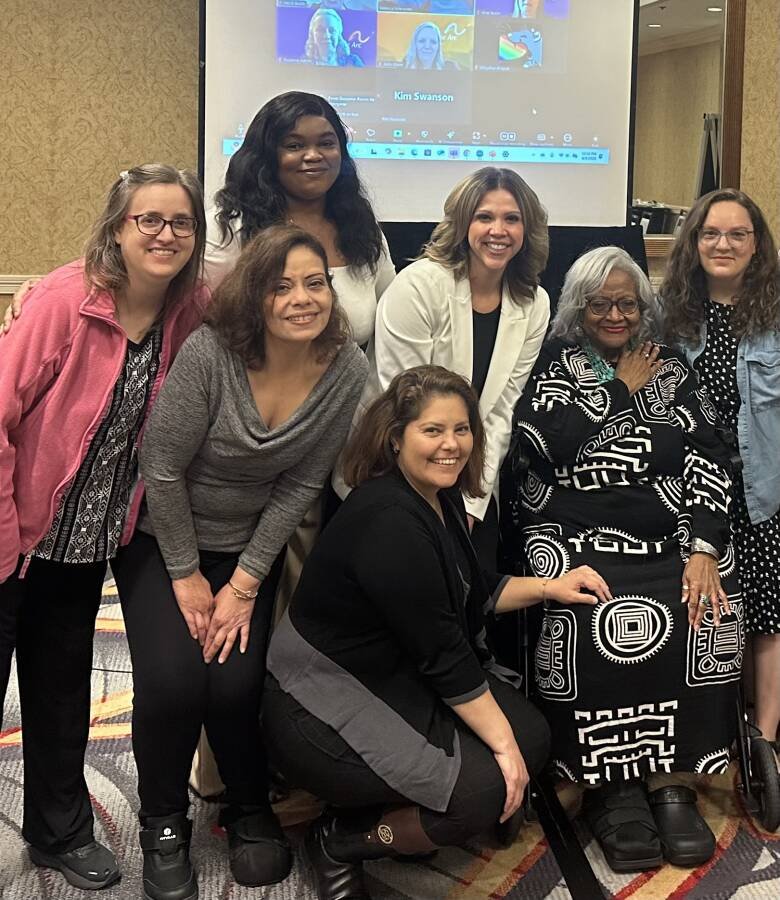 A group of nine women posing together for a photo at a conference or meeting room, with a projector screen behind them displaying a virtual meeting call.