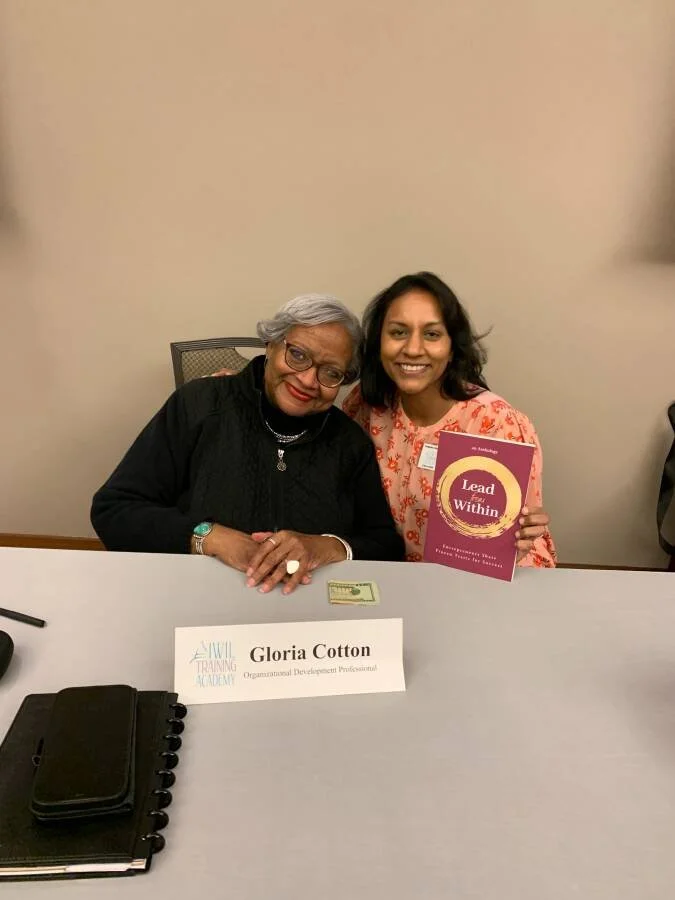 Two women. One older woman with gray hair and glasses, and one younger woman with dark hair and a pink patterned dress. They are sitting at a table with a nameplate that reads "Gloria Cotton, Organizational Development Professional", a notebook, and some papers. The younger woman is holding a pink book titled "Lead from Within" and both are smiling.