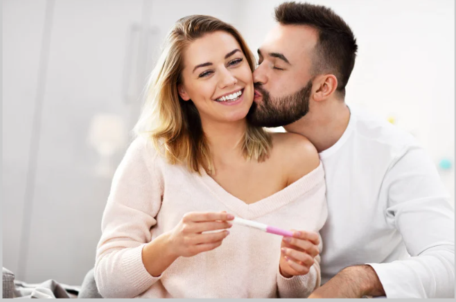 A man kisses a woman on the cheek while she smiles happily, holding a pregnancy test.