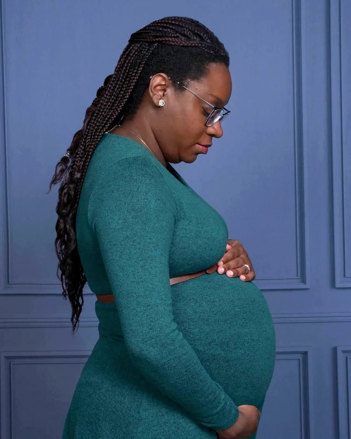 A pregnant woman with braids, glasses, earrings, and a wedding ring, wearing a teal dress, standing against a blue wall with decorative panels, looking down at her belly.