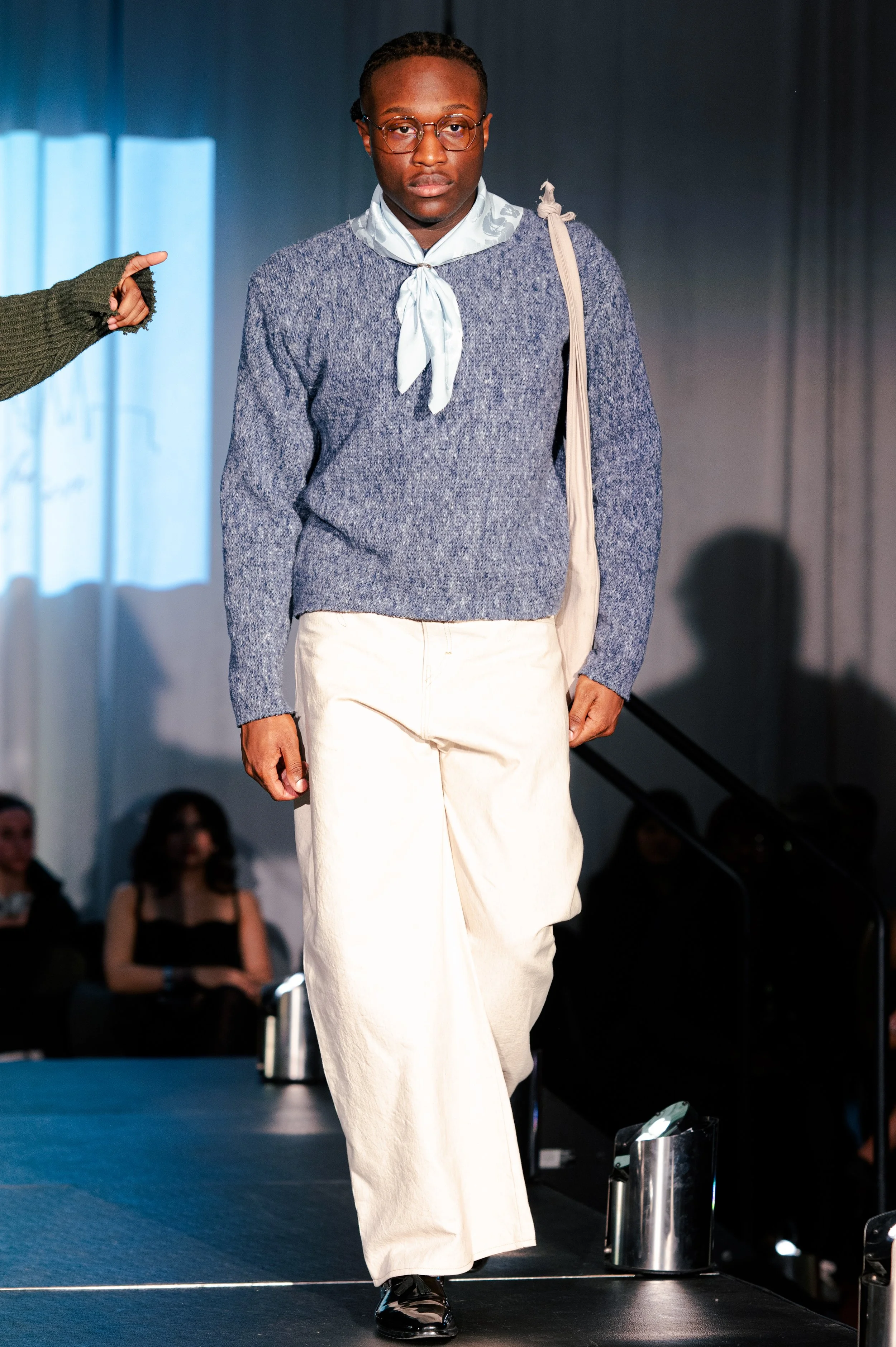 A male model walking on a runway during a fashion show, wearing a blue sweater, cream wide-leg pants, glasses, and carrying a bag over his shoulder.