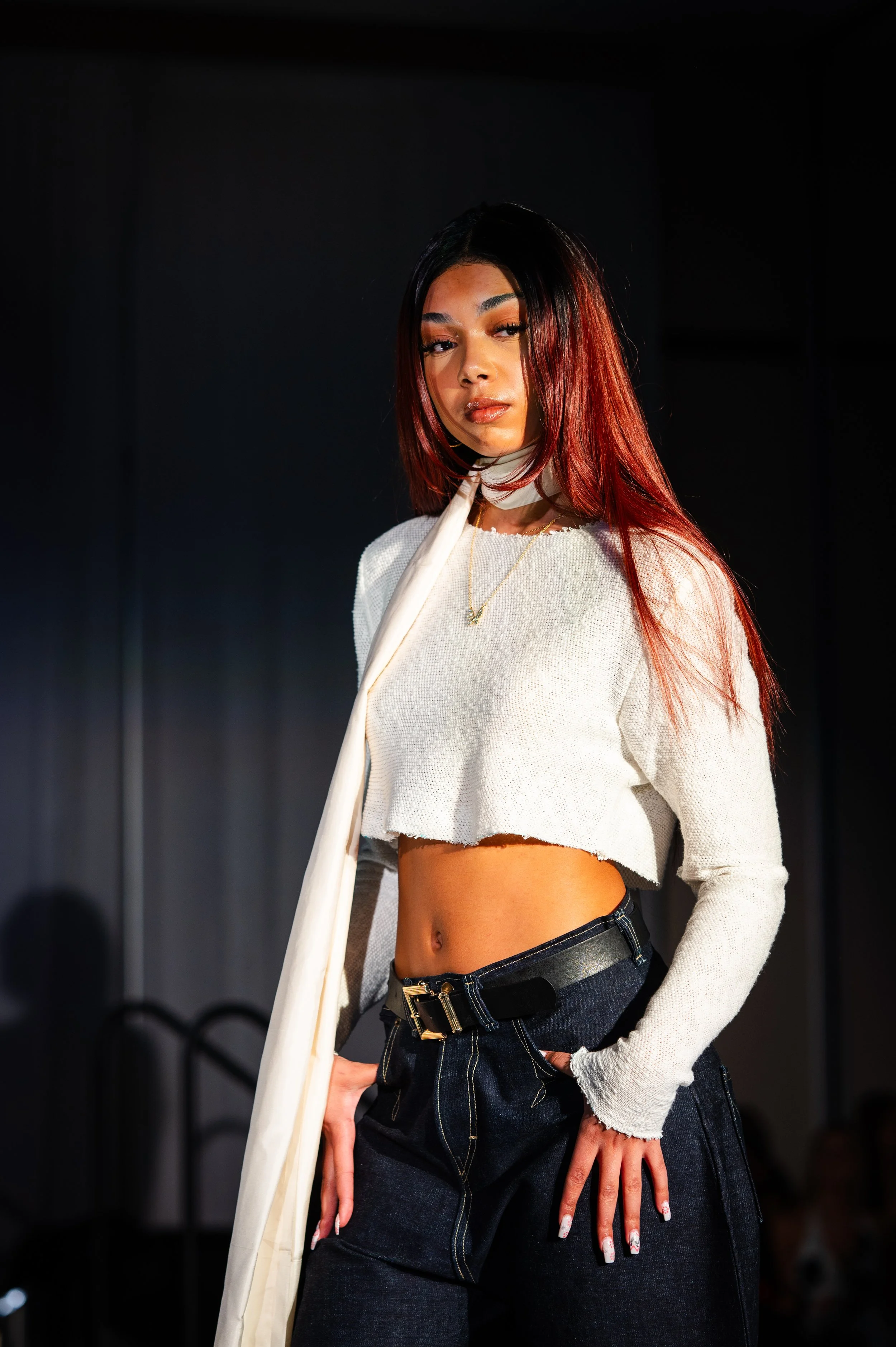 A young woman with long, dark hair featuring red tips, wearing a white cropped sweater and dark jeans with a belt, standing confidently indoors with a dark background.